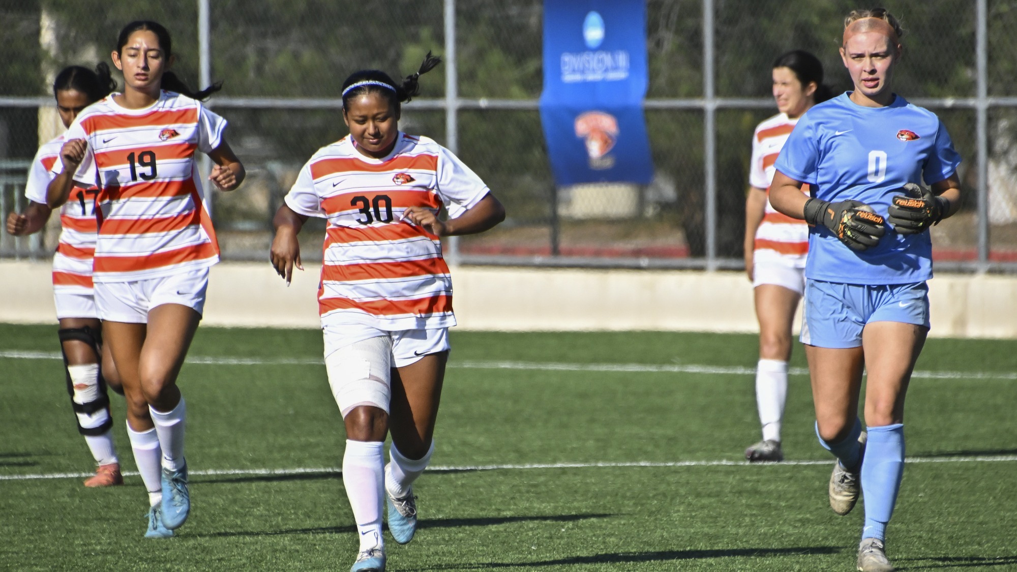 Haleigh Gardner, right wearing blue, Meher Banik, center, and Anusha Chatha run out onto the field towards the camera. The latter two wear orange and white horizontally-striped jerseys. 