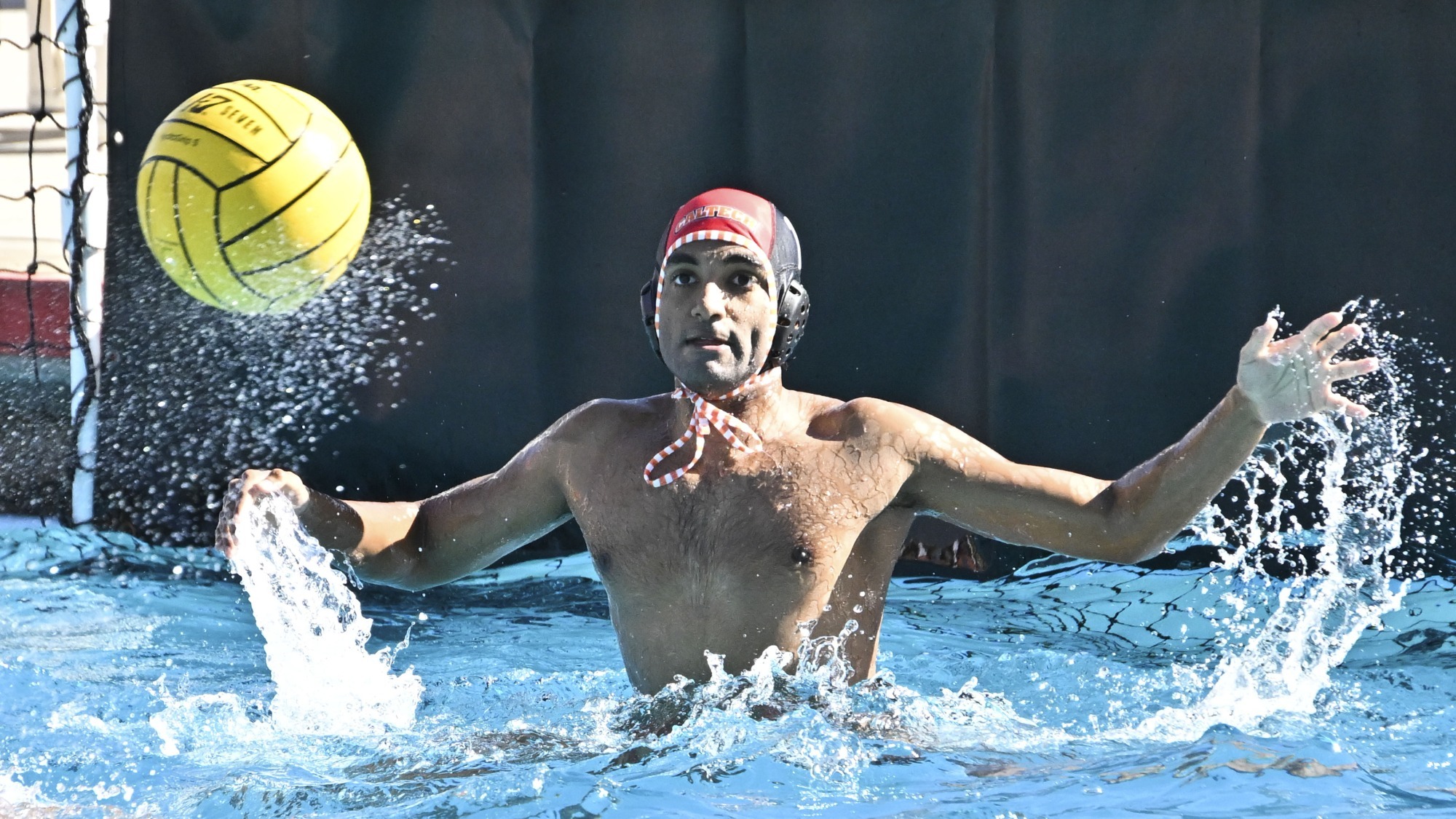 Sujit Iyer extends his left hand out of the water and is in the process of raising his right as he spreads his arms to make a save. His eyes are clearly focused on the ball in the top left of the frame which casts off droplets of water as a result of its rotation. 