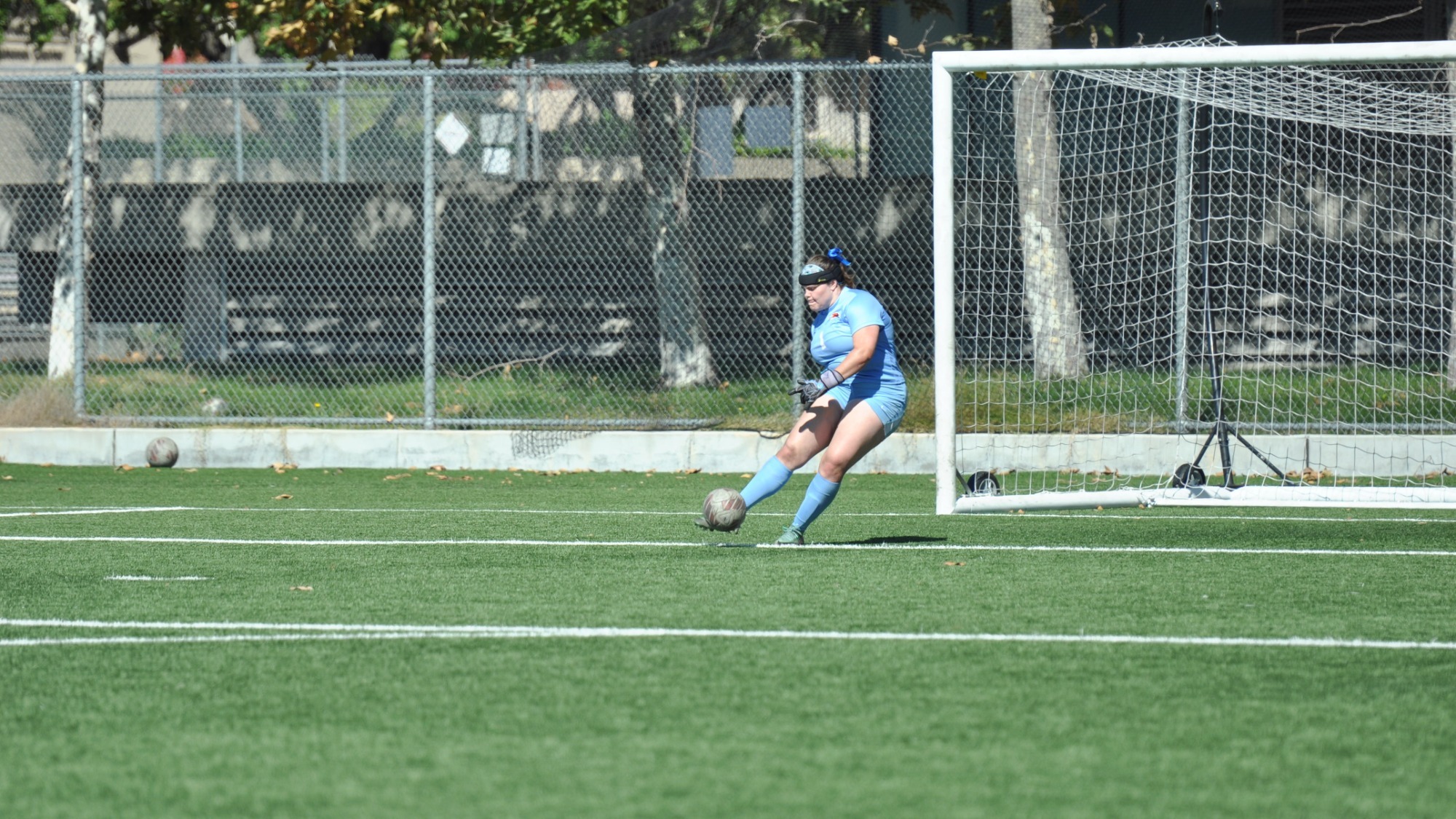 Sophia Steven, wearing an all powder-blue goalie kit, makes contact with the ball while executing a goal kick in front of the right post of the net. The ball has a faded red and grey coloration similar to BB8 from 