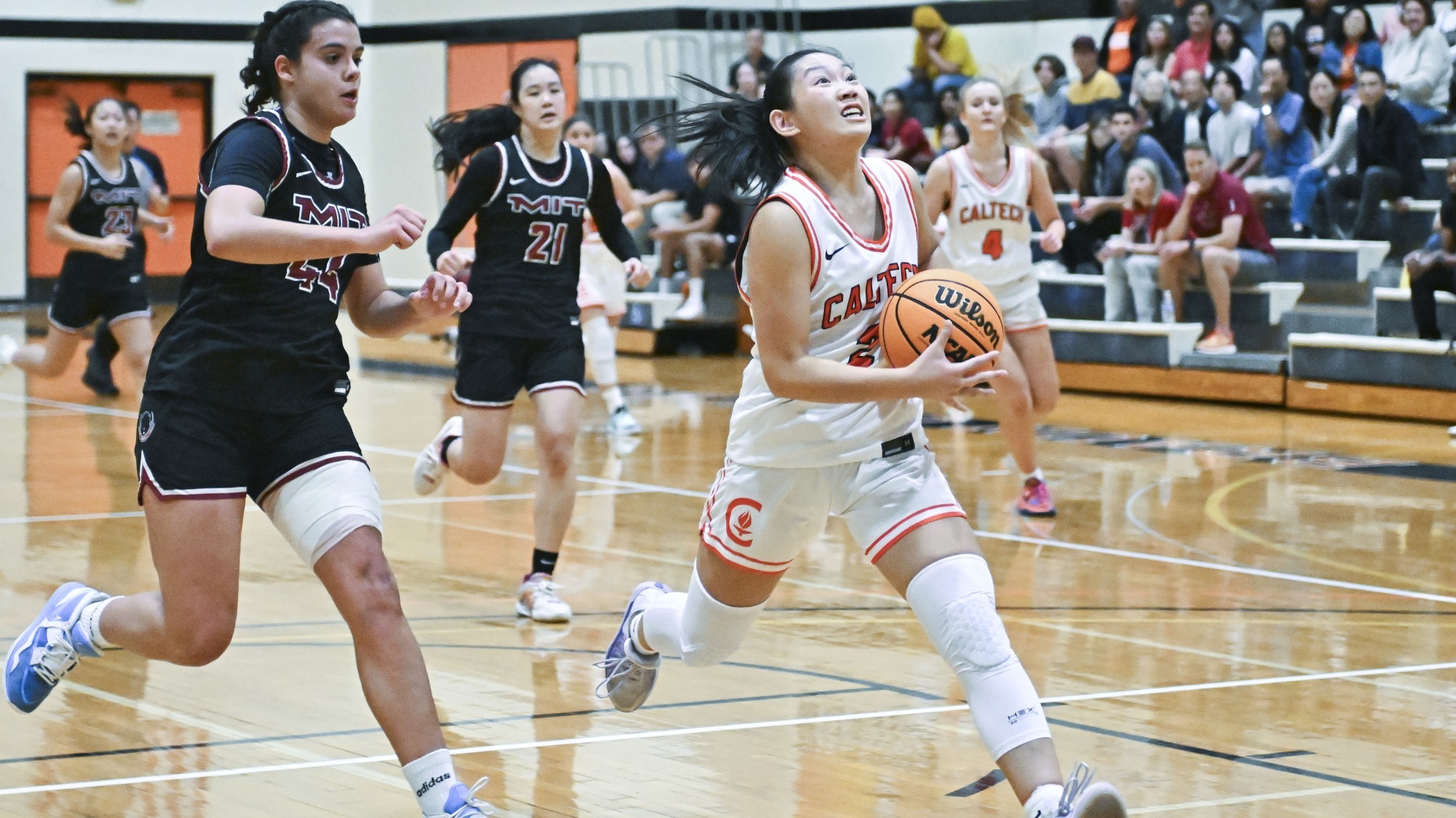 Kyra Phaychanpheng looks upward on the sprint as she prepares to go up for a layup against MIT. She holds the ball in both hands and is pursued by an MIT defender, 