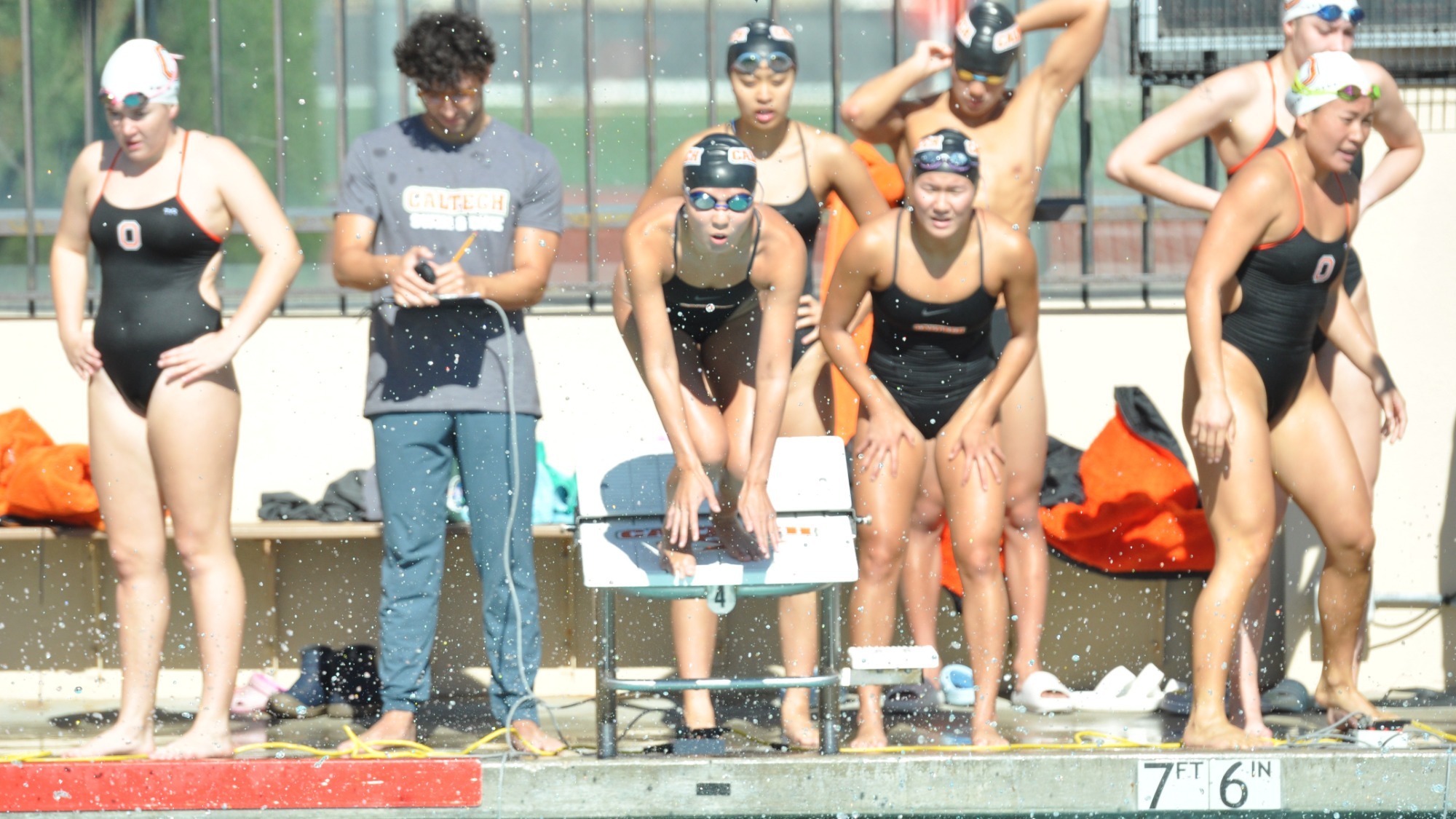 A member of a Caltech relay team crouches as she prepares to jump into the water for her leg of the relay. Two teammates stand behind her, cheering. 