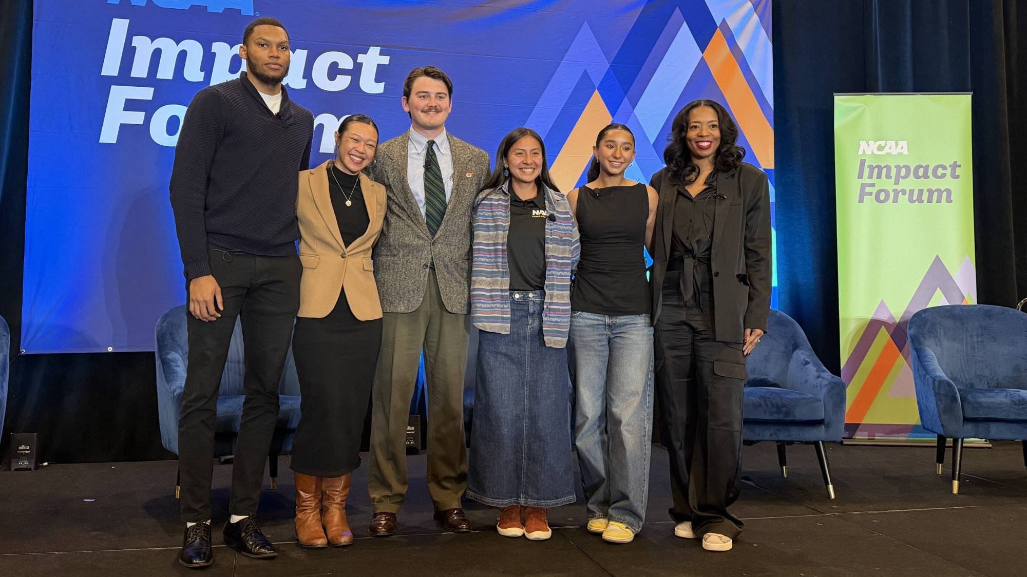 Aidan Hamner stands in a full suit and tie on the stage of an NCAA Impact Forum event, judging by the branding in the back ground of the photo. Hamner stands third from the left in a group of six people. 