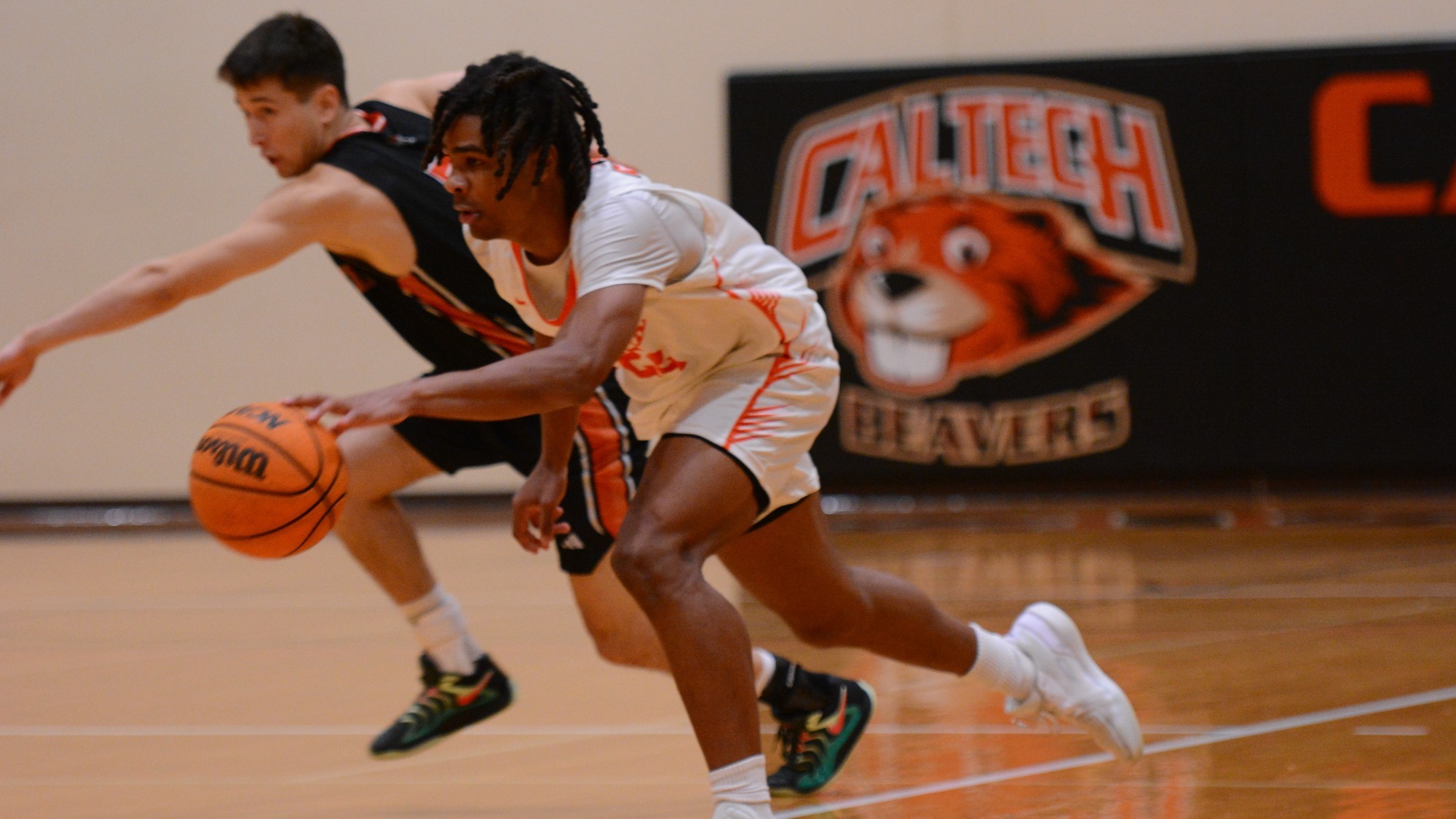 Chase Williamson Drives to the basket in a white jersey with Orange numbers. An Occidental defender in a black jersey races towards him from behind, apparently having just been beaten on the dribble. 