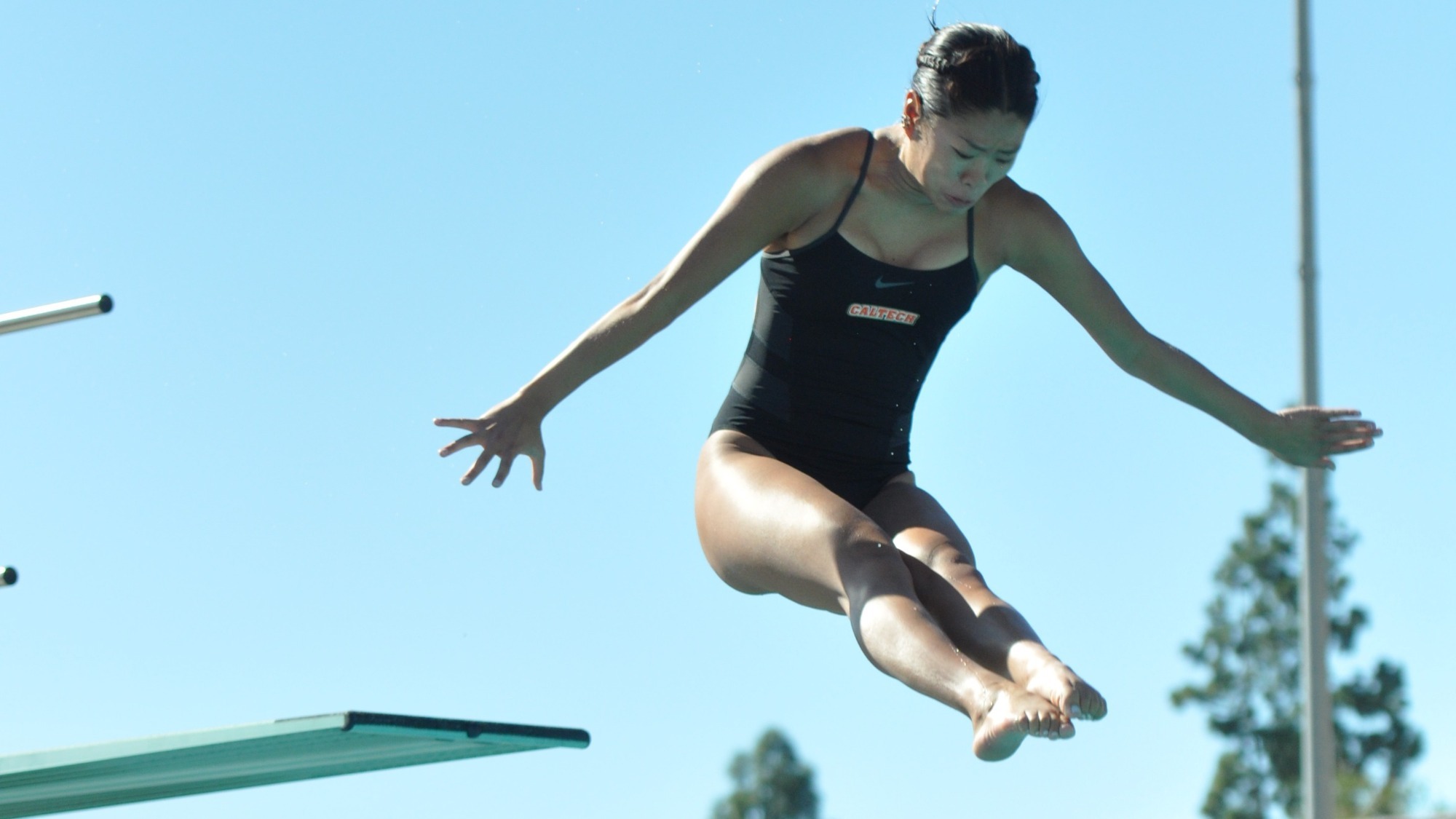 Jana Woo looks down at the water with her head above the diving board and her feet below it as she unfolds from the pike position. She wears a black swimsuit with 