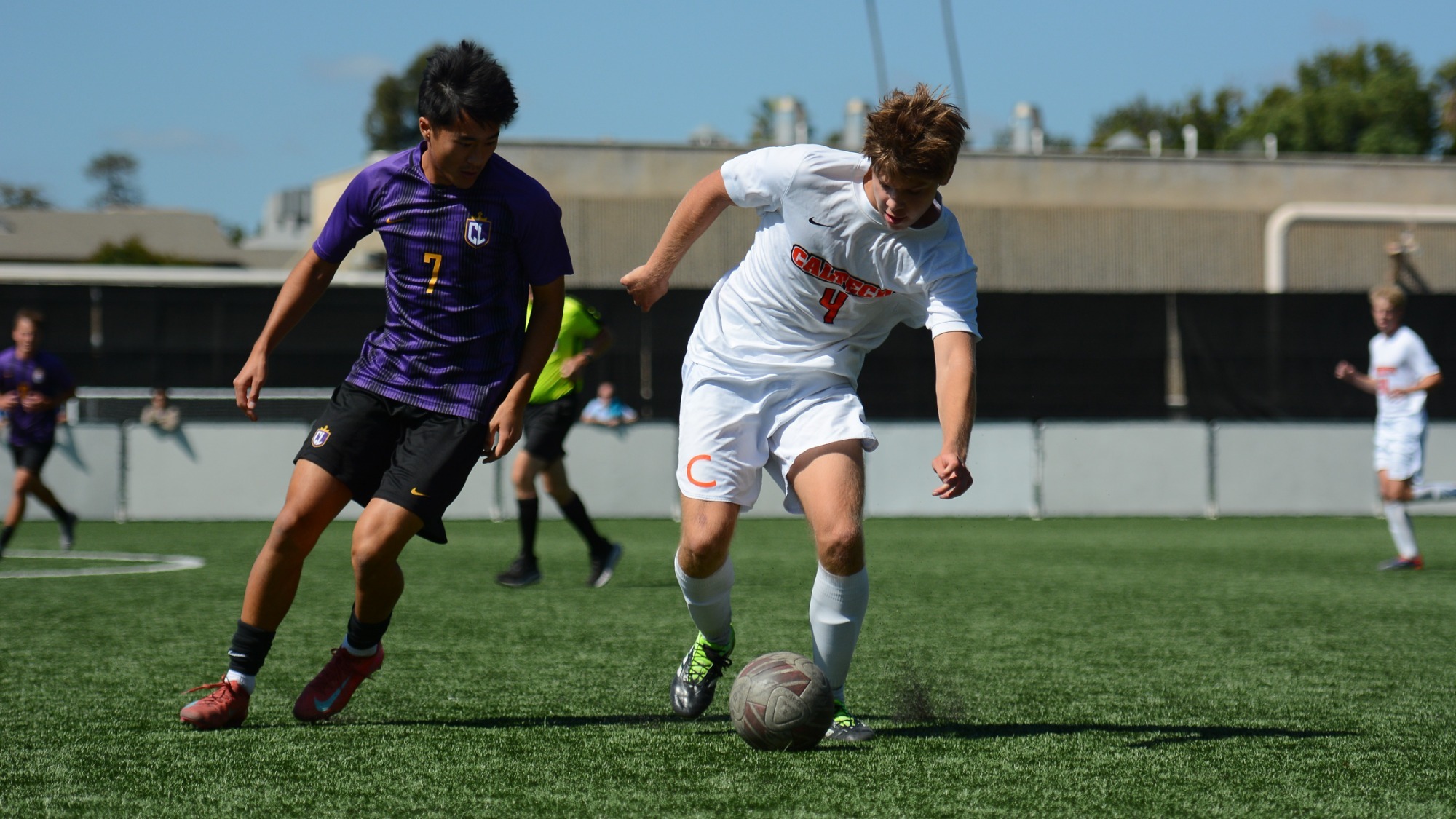 Aritz Schube in a white jersey with an orange number 4 on the front dribbles, defended on his right (left of frame) by a Cal Lu defender in a purple jersey with faded black vertical stripes and a gold number 7. 