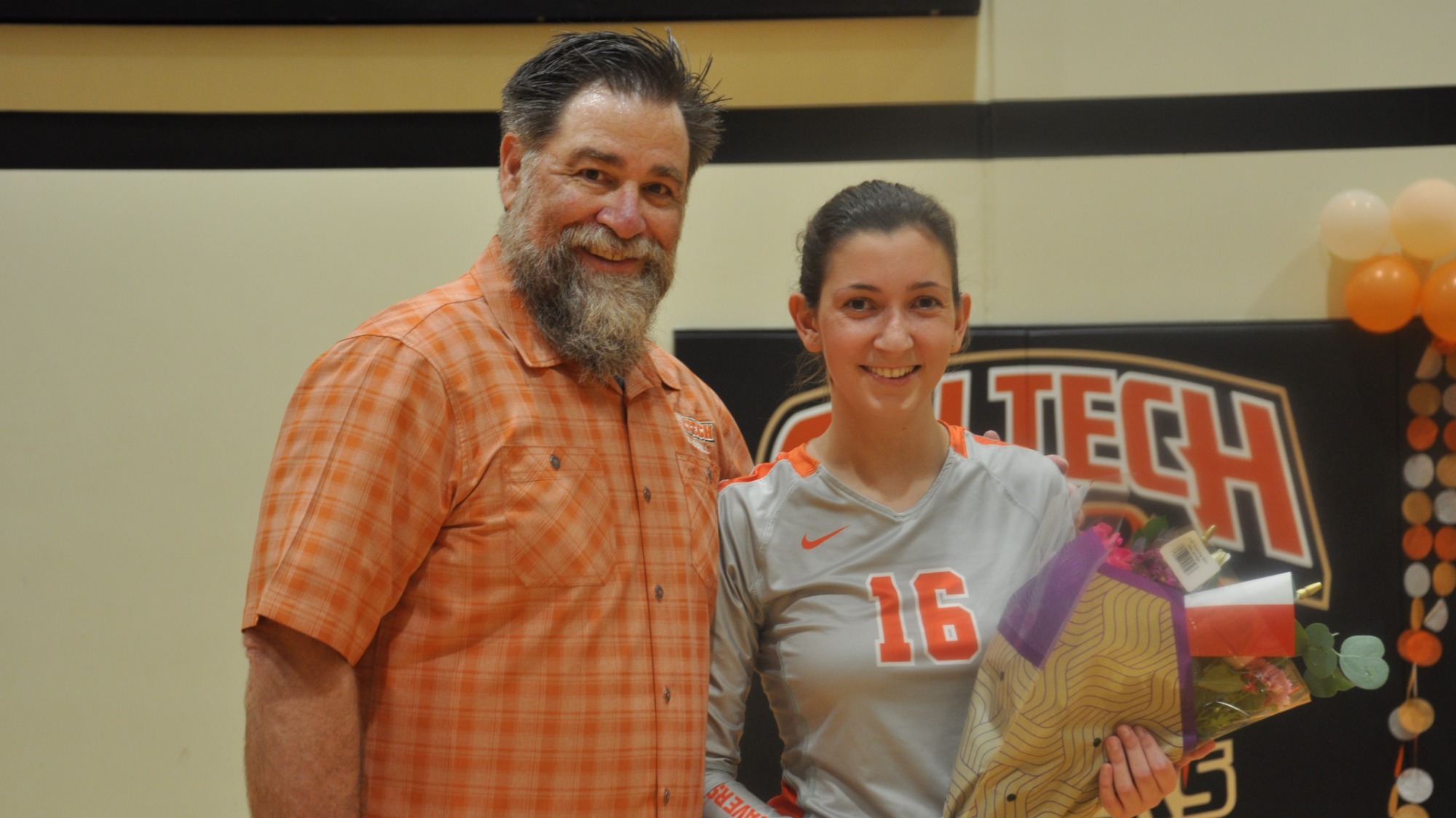 Guilia Murgia stands to Tom Gardner's left in her grey jersey with orange numbers holding a bouquet 