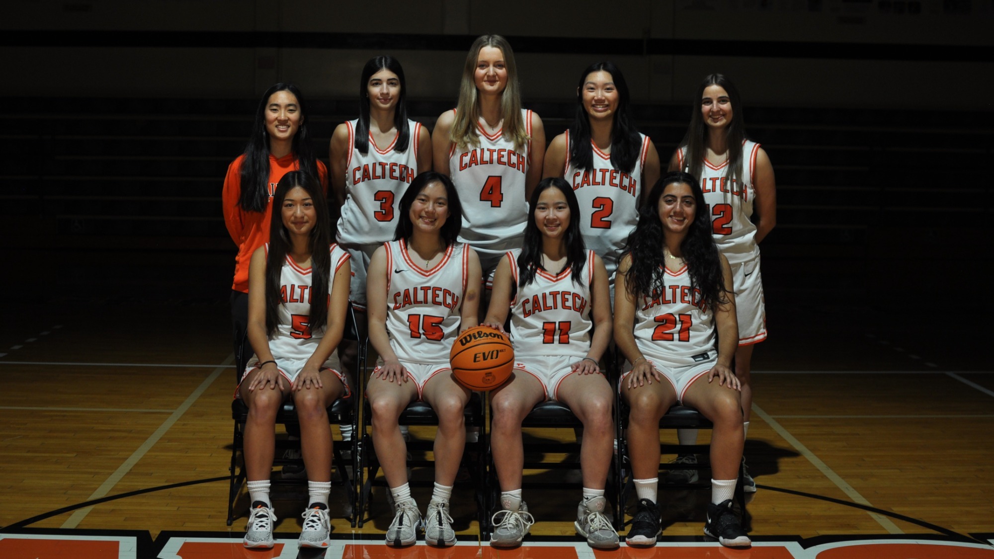 The women's basketball team poses for a team photo in a darkened Braun Gymnasium. The front four players sit while the back four players and coach Quan stand. The team is in a spotlight wearing their white uniforms with orange lettering