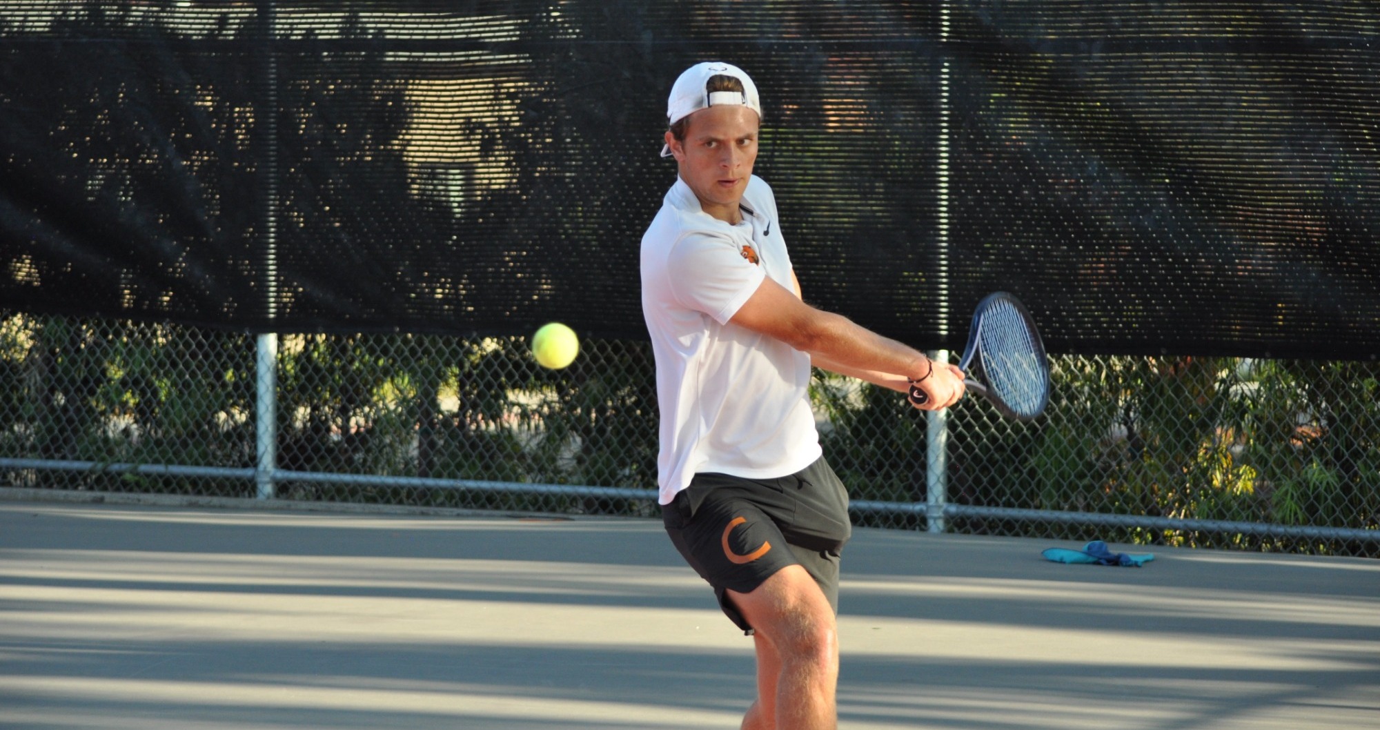 Constantin Cedillo-Vayson de Pradenne prepares to hit a two-handed backhand from the short side of the court and the left side of his body. Wearing a backwards white baseball cap and a white tee, his eyes focus on the oncoming ball while his arms reach back to swing. 