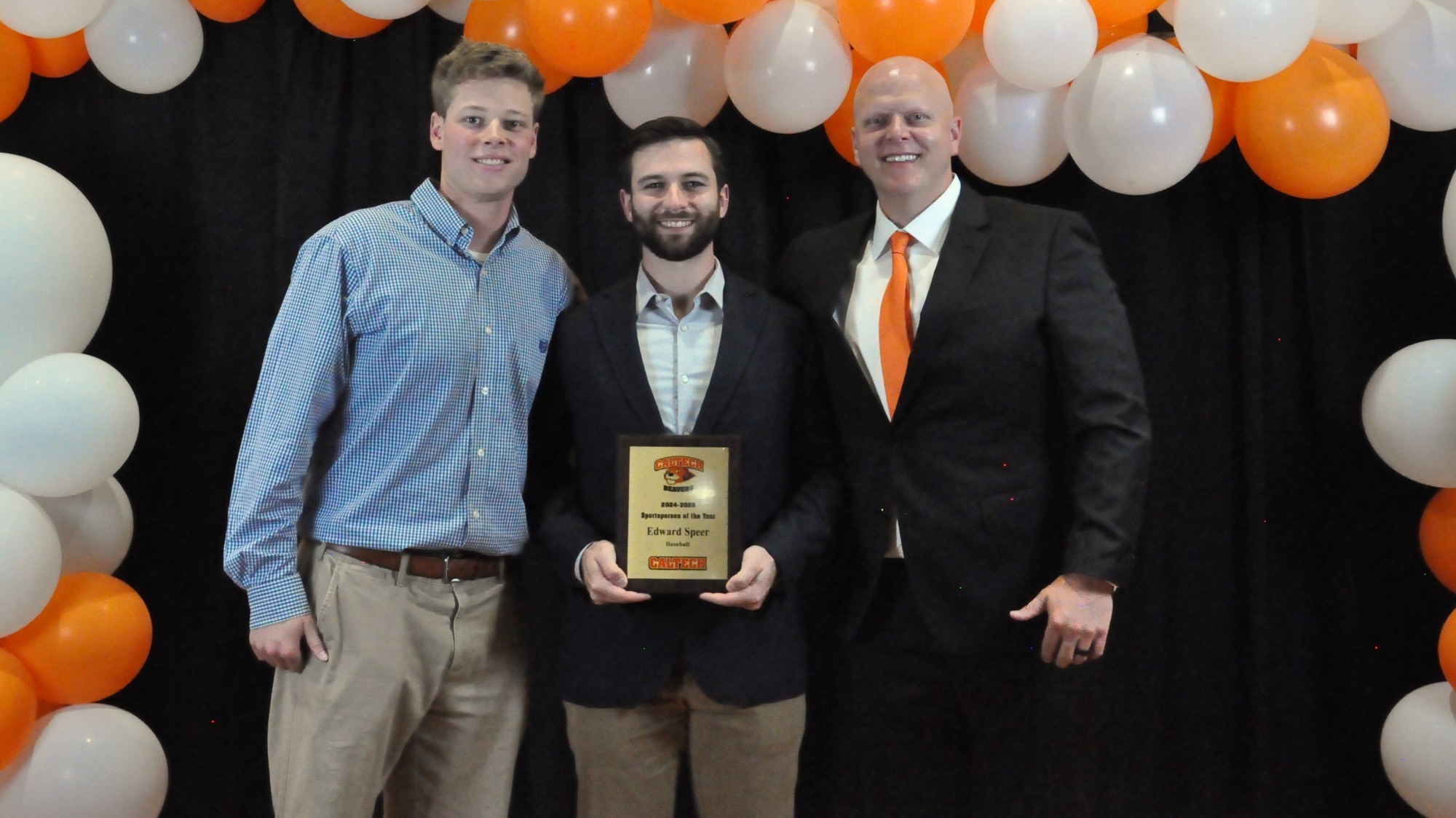 Coaches Frank Craska (left, blue shirt) and Kevin Whitehead (right, suit and orange tie, white shirt) stand with catcher Edward Speer (middle) for a photo. Speer holds a plaque commemorating his designation of Male Sportsperson of the year at Caltech. 