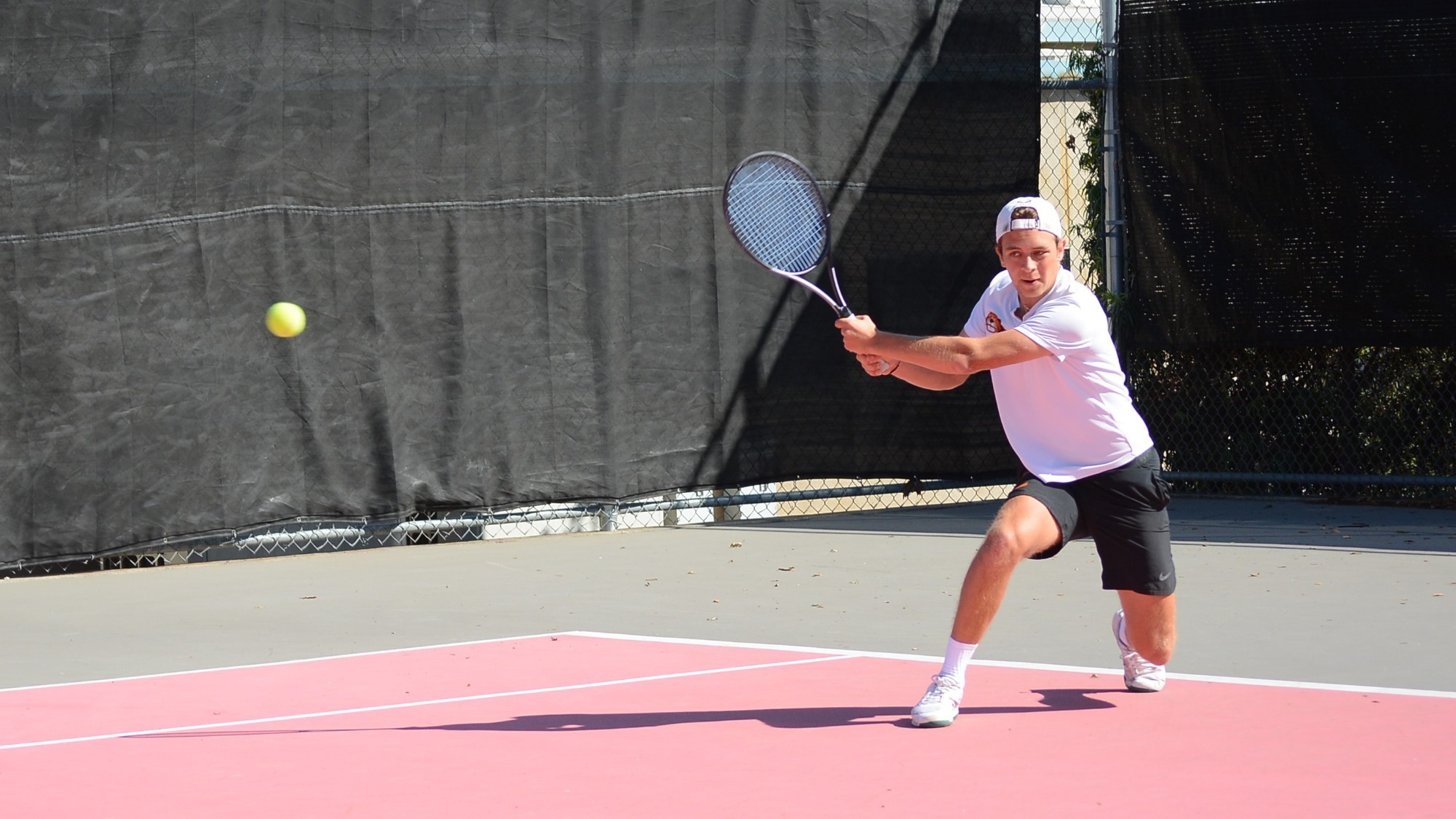 Cedillo-Vayson de Pradenne watches the ball approach as he prepares a two-handed backhand swing. He is wearing a white T-shirt and a backwards white baseball cap 