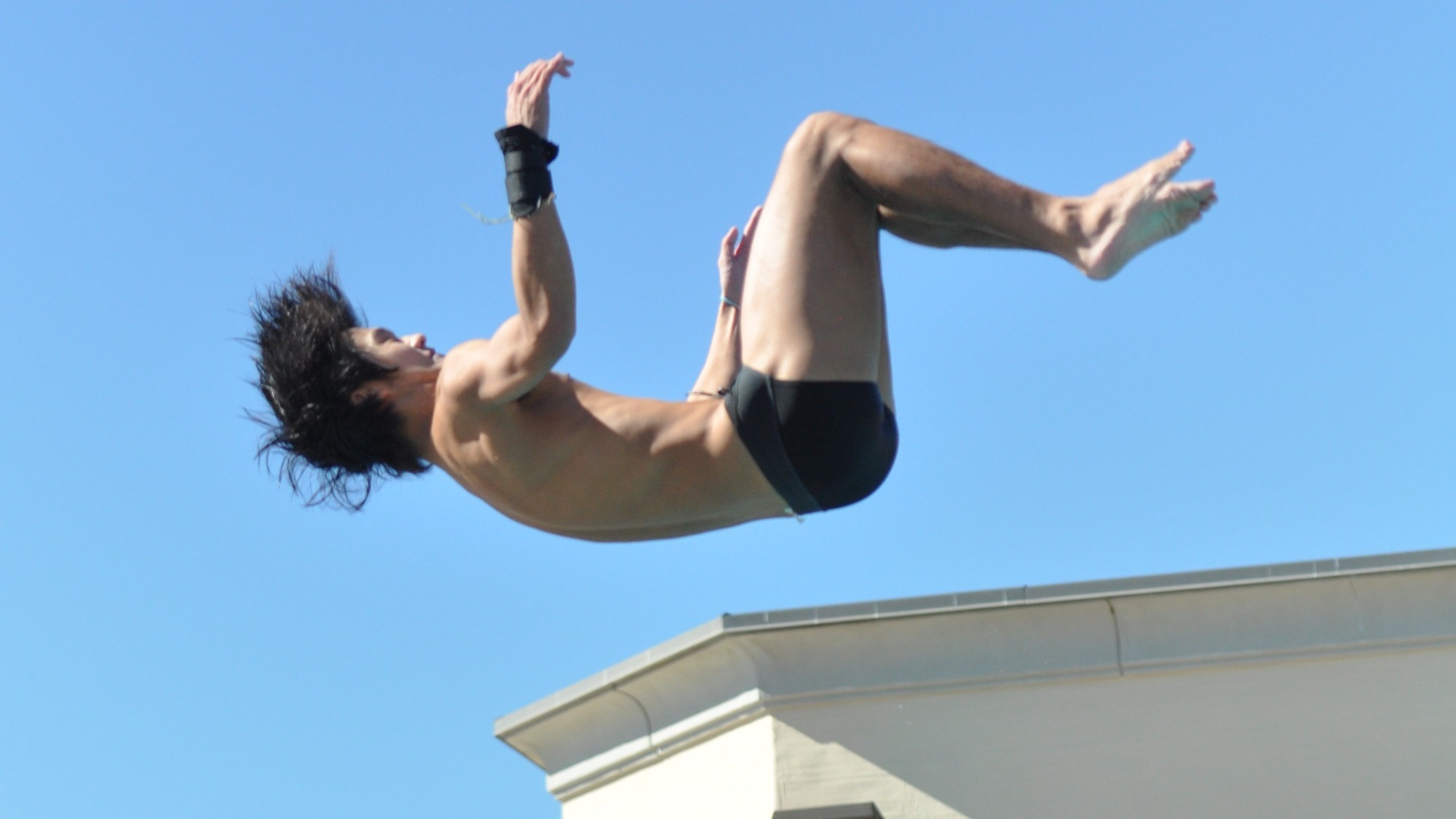 Anthony Wang's hair ripples as he initiates a backflip above the height of the diving board with his back parallel to the surface of the water. 