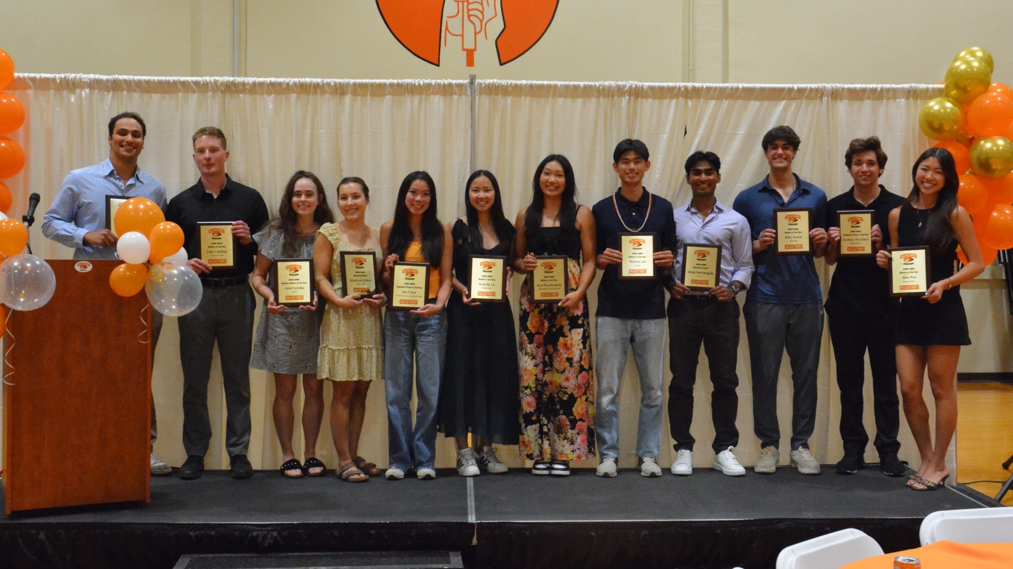 The Award-winners of the 2025-26 Student Athlete Celebration stand together onstage in business-casual attire with their award plaques. 
