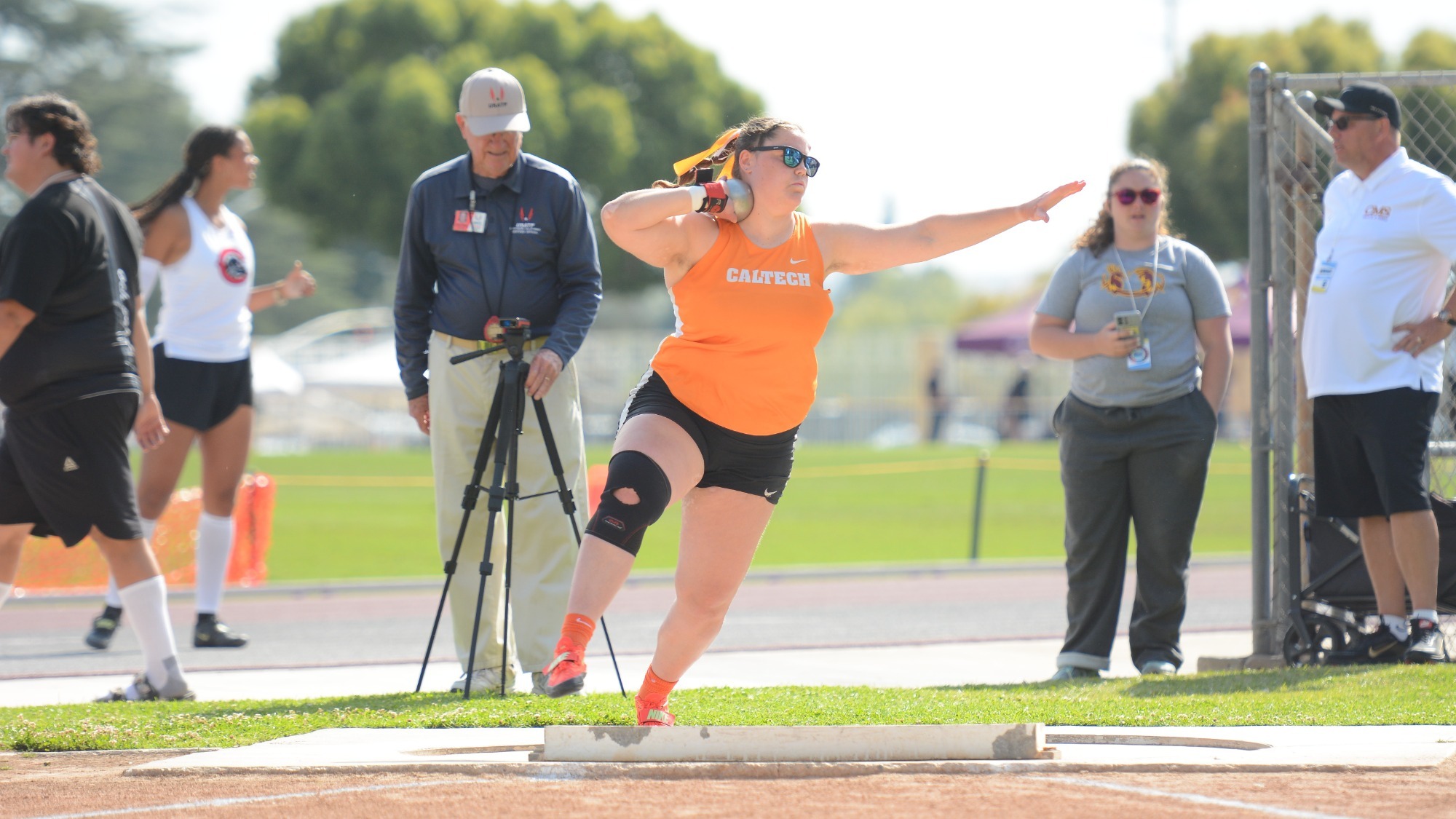 Sophia Steven, wearing and orange top and sunglasses, holds the shot against her neck as she initiates her toss. 