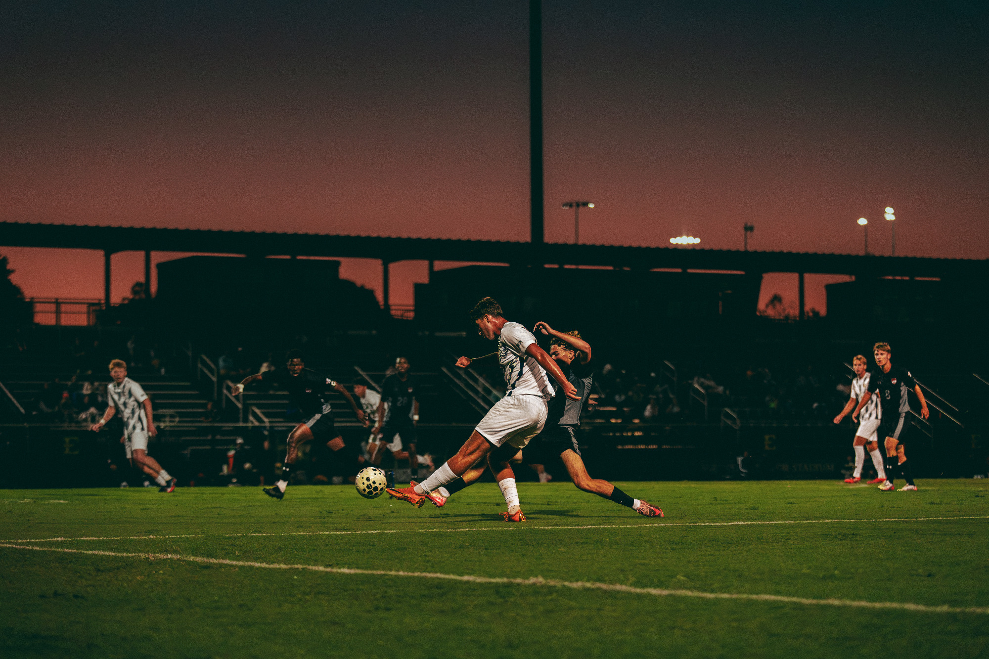 Gavin Dick fires a left-footed shot on goal from a tight angle along the end line during a soccer match against Gardner-Webb. The grandstands are in silhouette in the background as the sun sets behind them.