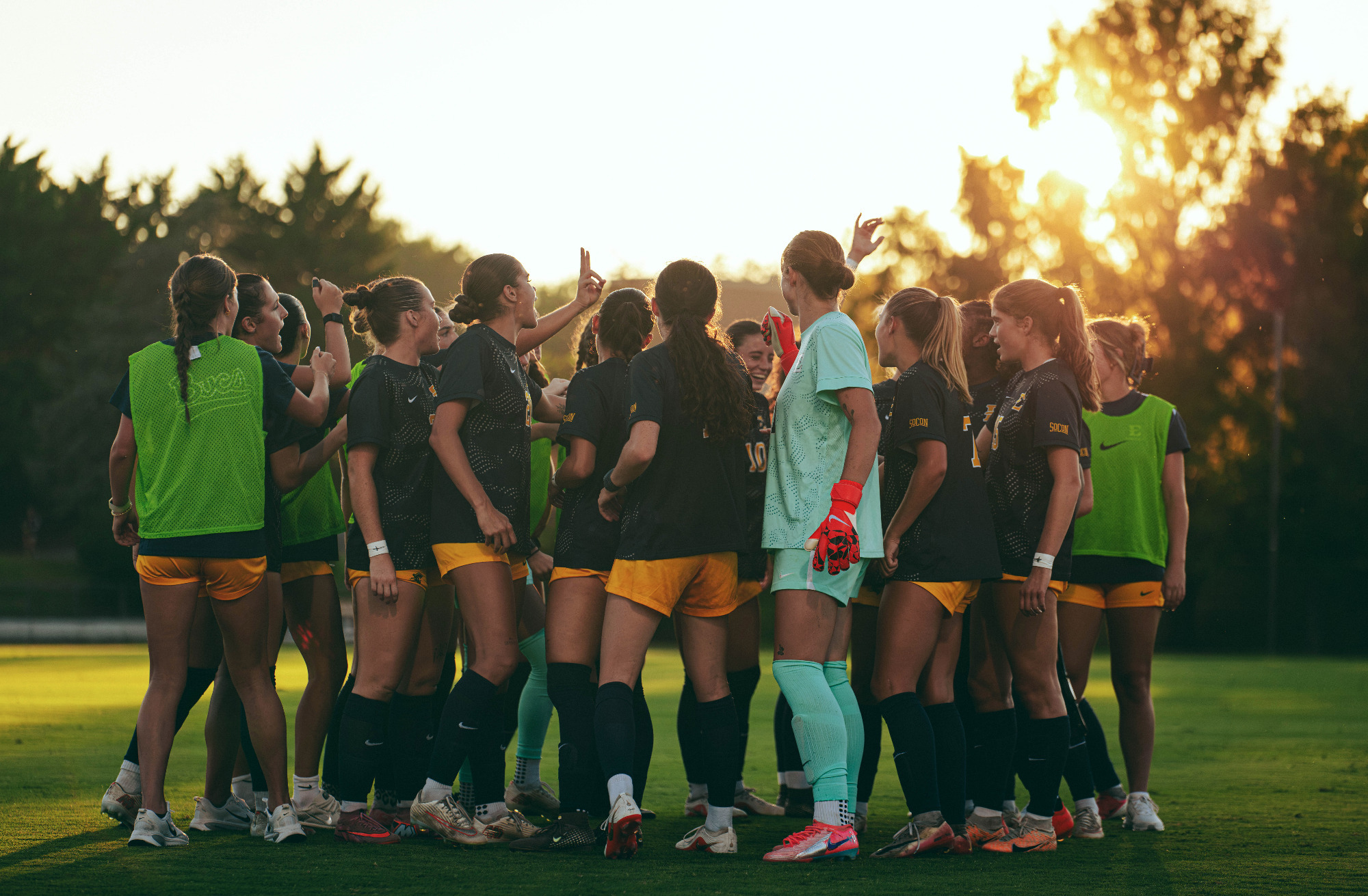 The ETSU women's soccer team huddles before a soccer match against Radford. The setting sun casts them in silhouette, aided by the dark blue uniforms of the outfield players.
