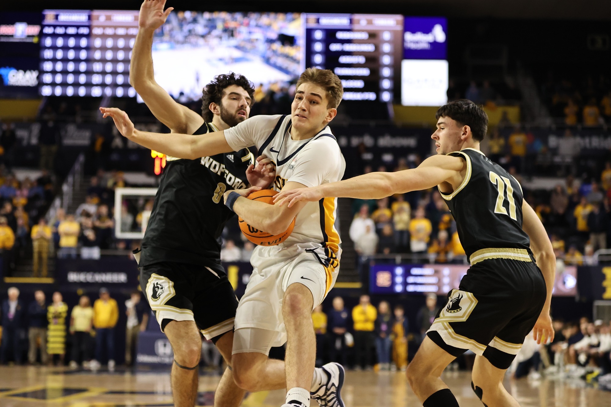 Blake Barkley, in ETSU's white home uniform, fights to drive toward the basket between two Wofford defenders wearing their black and gold road uniforms.