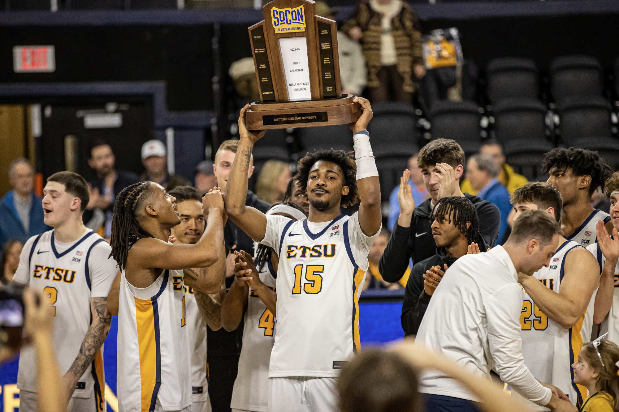 Cam Morris, still wearing his white basketball uniform, lifts the SoCon championship trophy over his head in front of his celebrating teammates. The trophy is made of wood with glass and brass elements.