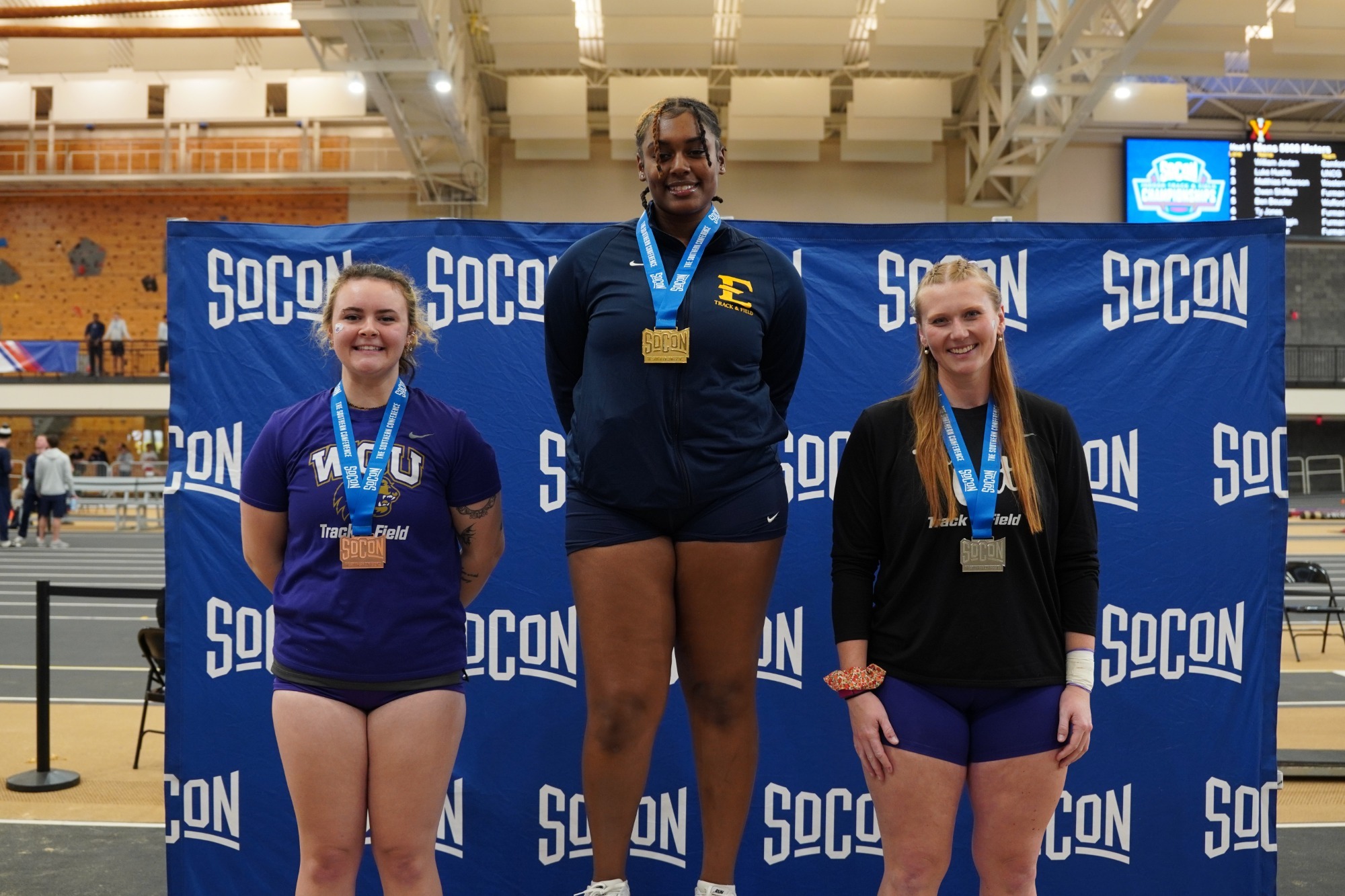 Tyla Nicholson stands on the top step of the podium wearing a gold medal over her dark blue ETSU track jacket. The silver medalist and bronze medalists, both from Western Carolina, stand either side of her in their purple and black team colors.