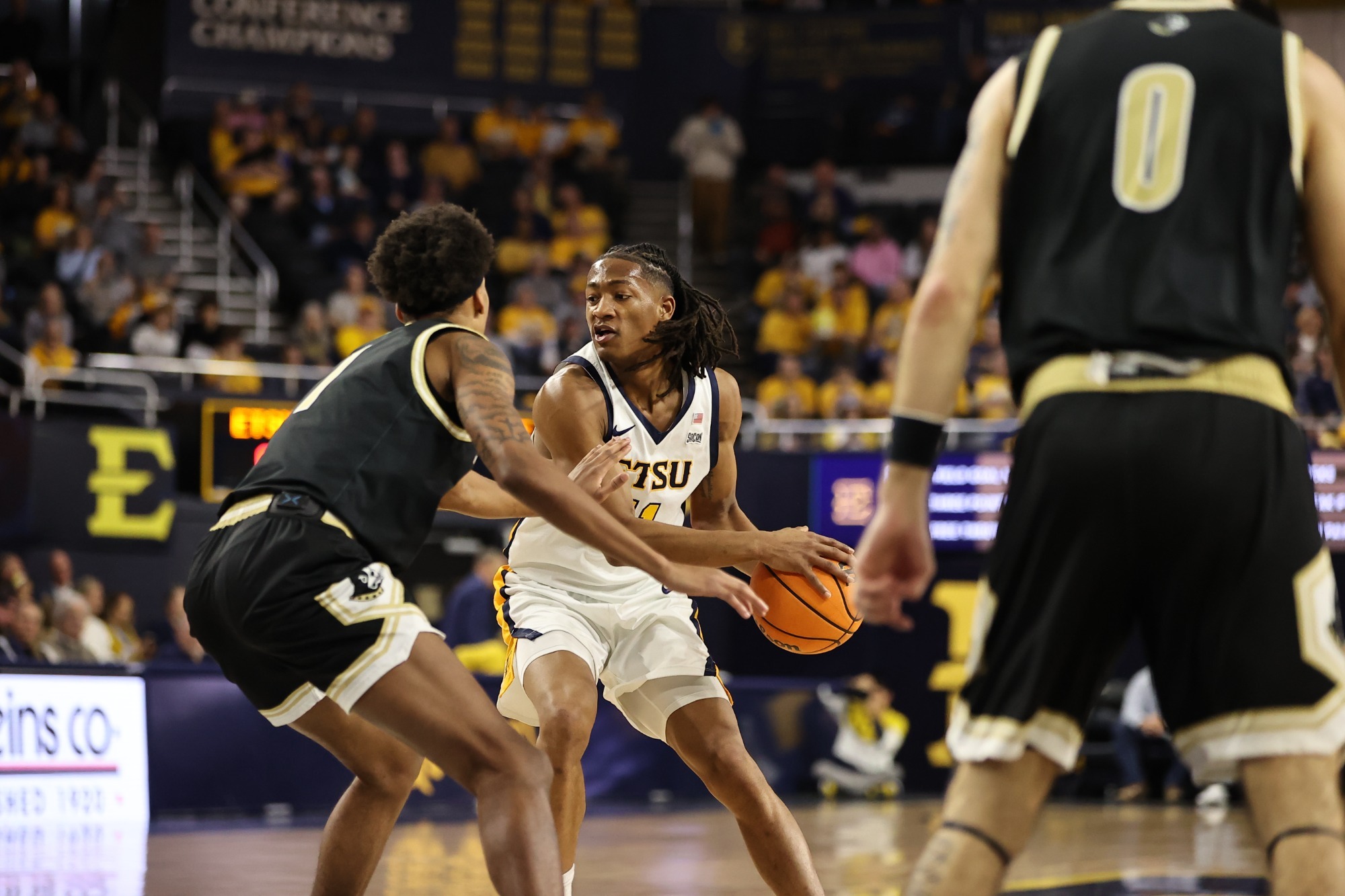 Brian Taylor, holding an orange basketball off the left hip of his white uniform, looks to drive through two Wofford defenders in their black and gold road jerseys