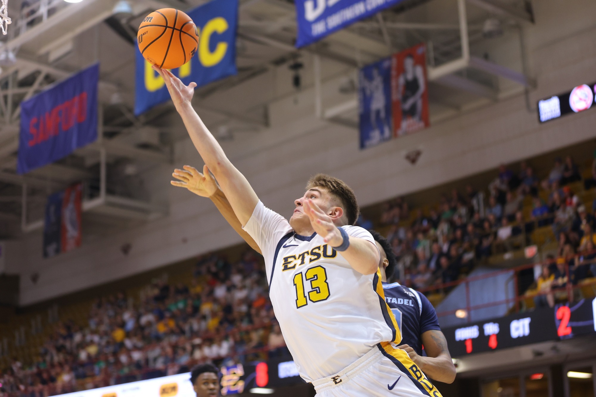 Number 13 Blake Barkley, in his white basketball uniform, reaches out with his right hand for a hook shot beneath the basket.