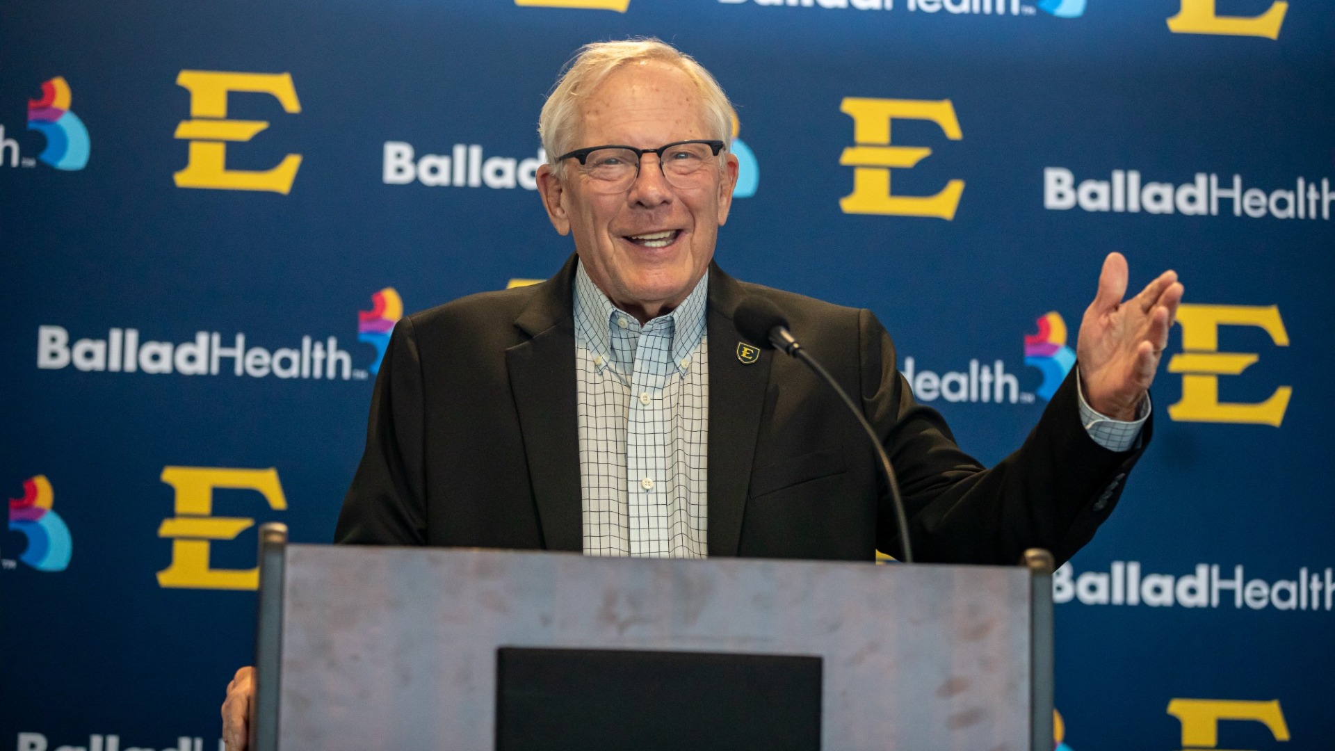 Dr. Richard Sander at podium in a black suit and striped button up shirt. He is smiling and raising his left hand. Backdrop has the ETSU E logo and the Ballad Health logo on a blue background.