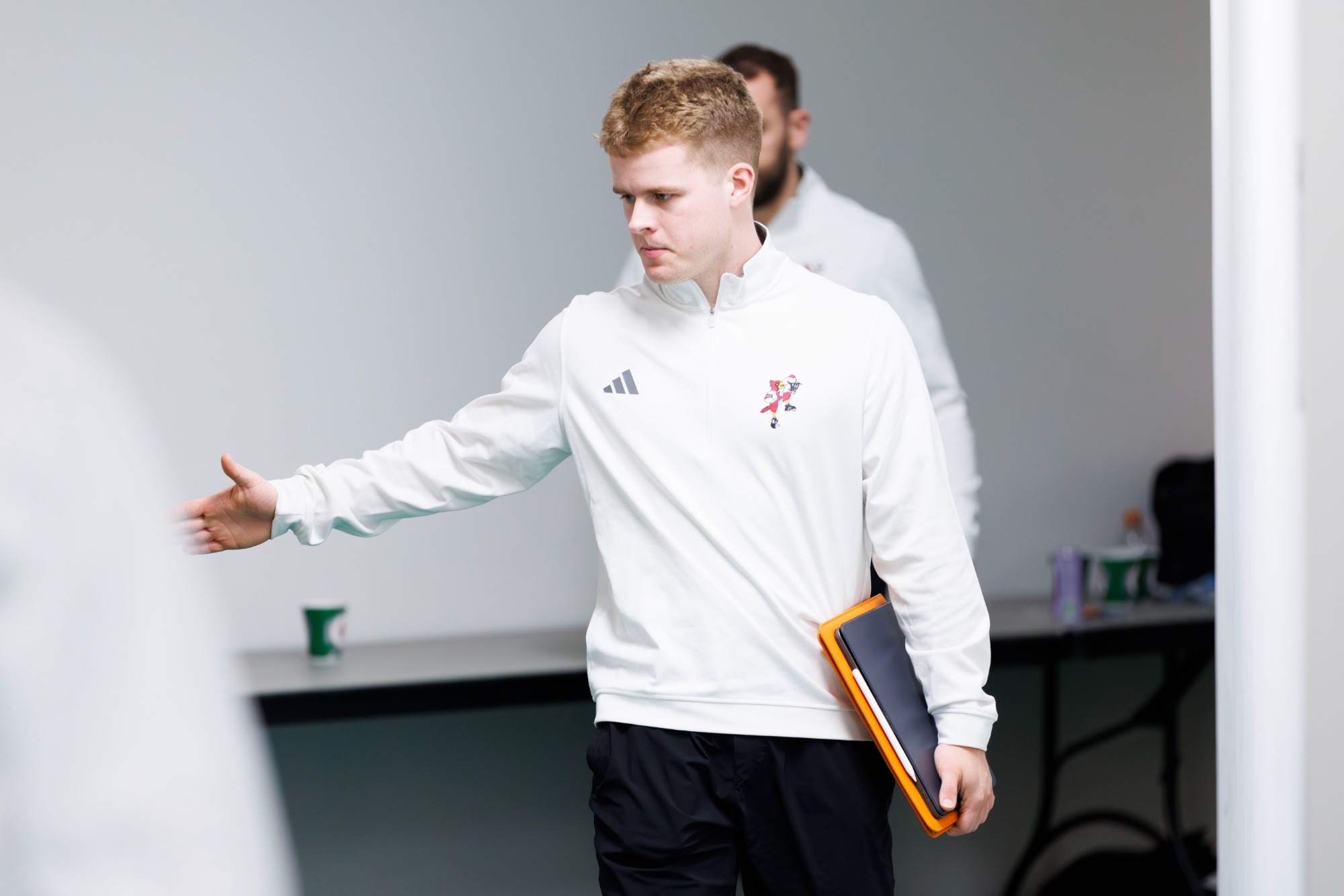 Graham Sandefur, in a white quarter zip and black trousers, extends his right hand to an unseen player outside the Louisville locker room