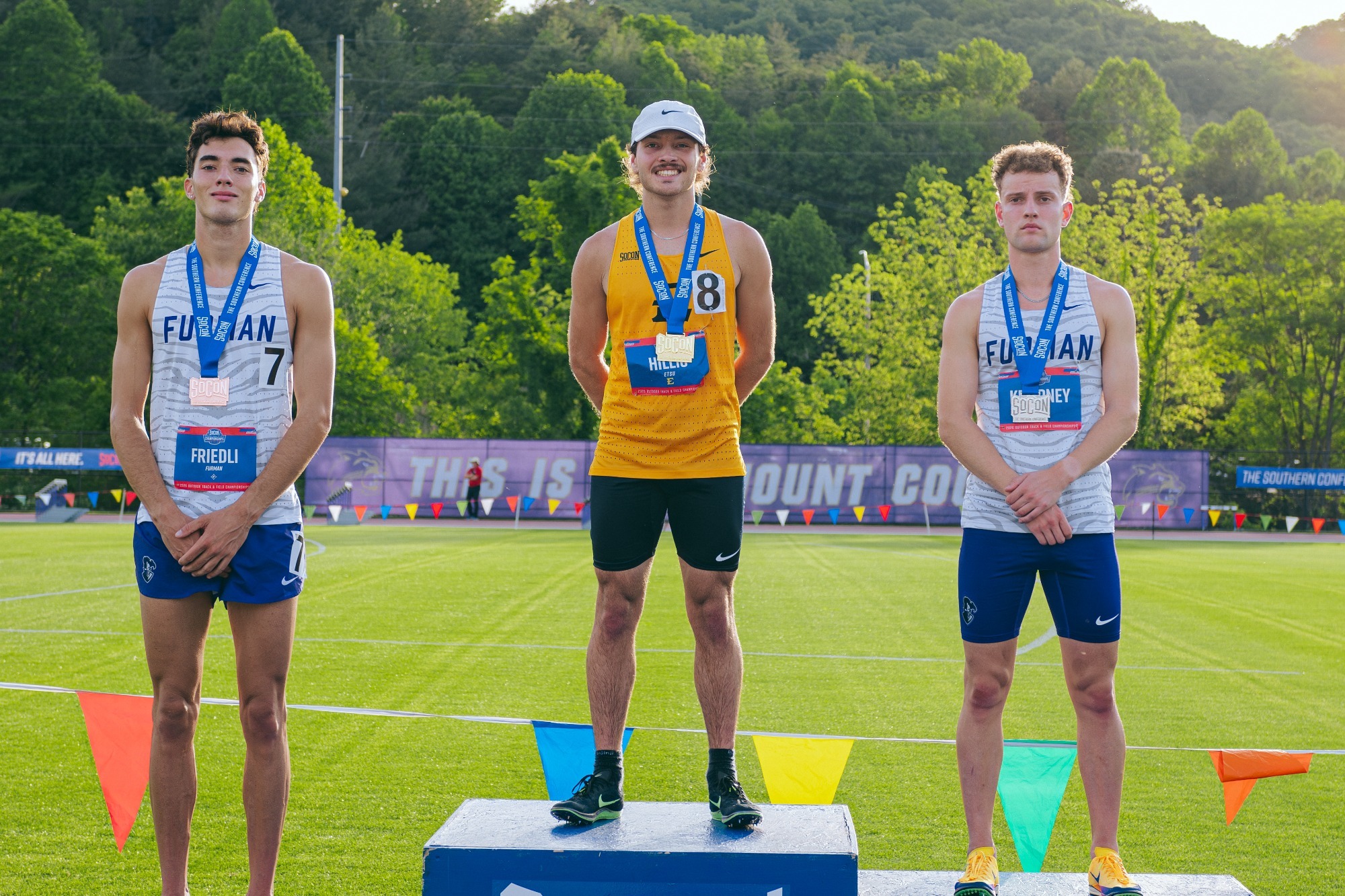 Nate Hillis stands on the center step of a podium with a gold medal from his 3000m steeplechase victory. Either side of him stands a runner from Furman wearing silver and bronze medals, respectively.