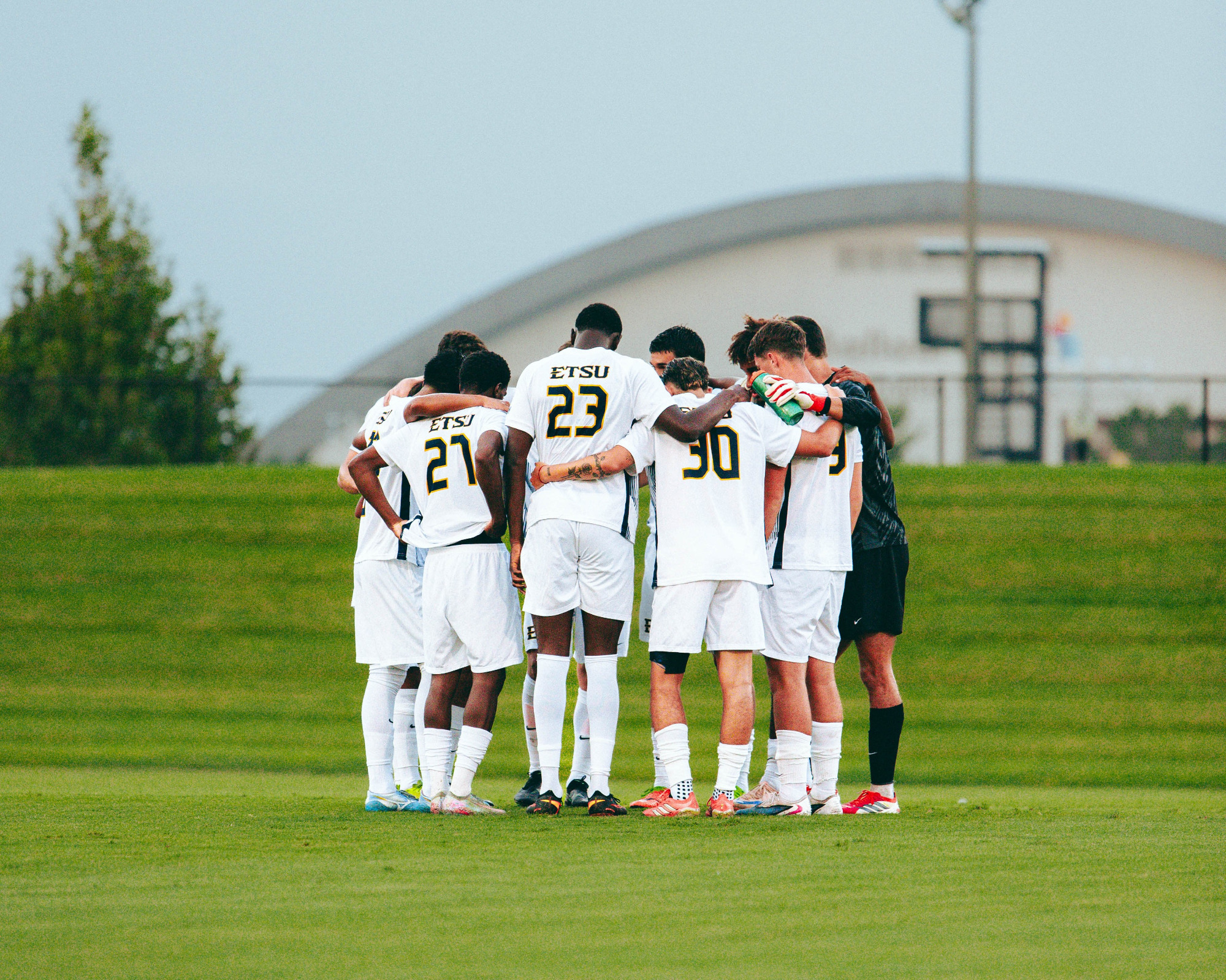 The ETSU men's soccer team's starting lineup huddles on the field in their white uniforms before a home game.