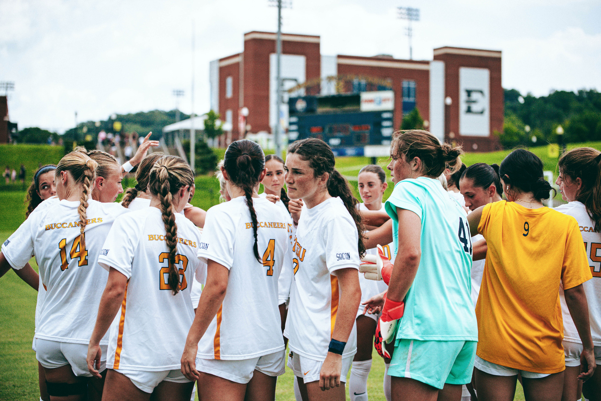 The ETSU women's soccer team huddles before their exhibition with USC Upstate. The players are in their white soccer uniforms except for Addison Yelton in her teal goalkeeper's kit and Bailey Coston in her gold warm-up jersey.
