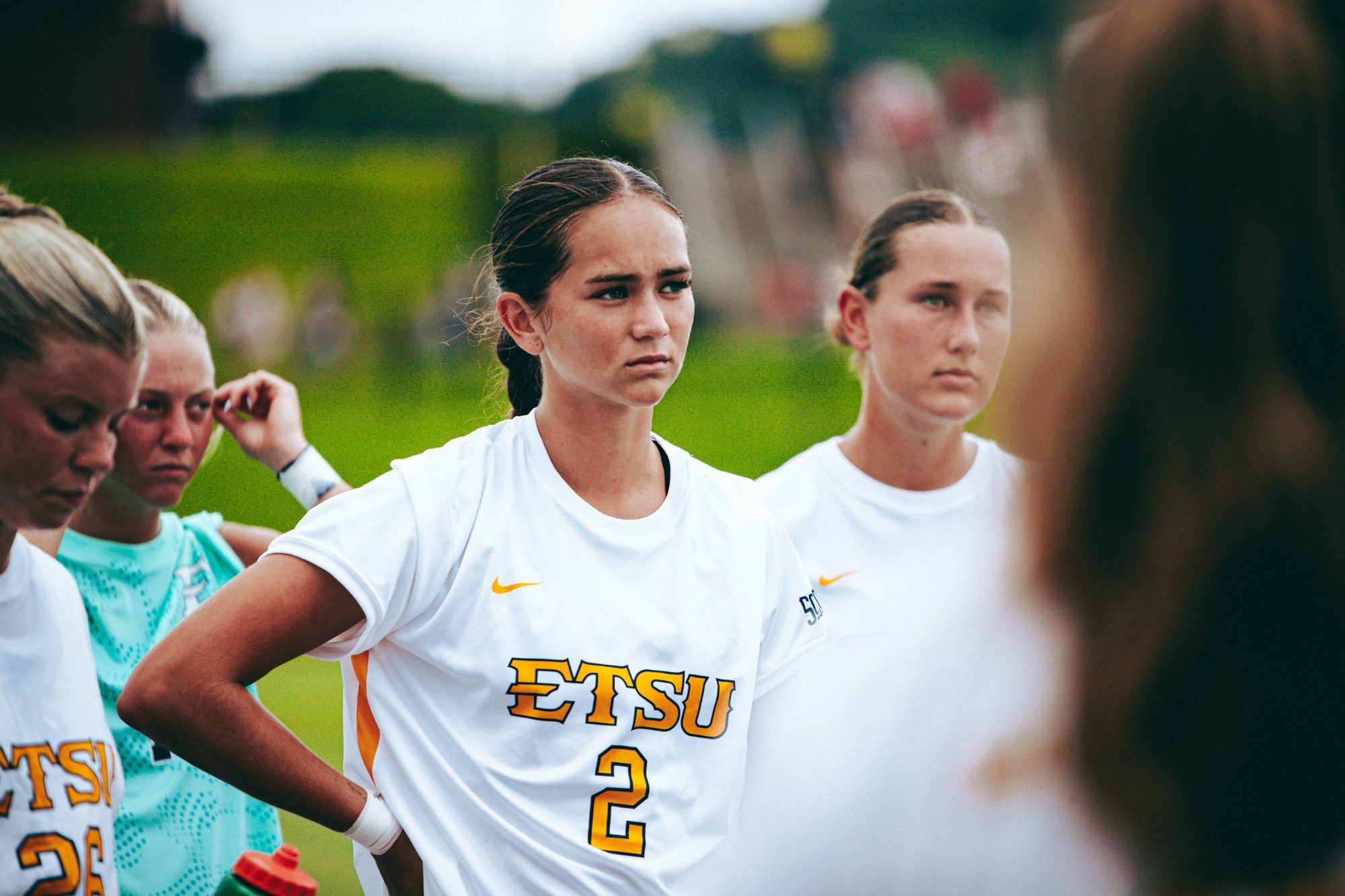 Alex Beck stands in the huddle with her teammates with her right hand braced against her hip, listening intently to instruction from a coach just out of frame on the right.
