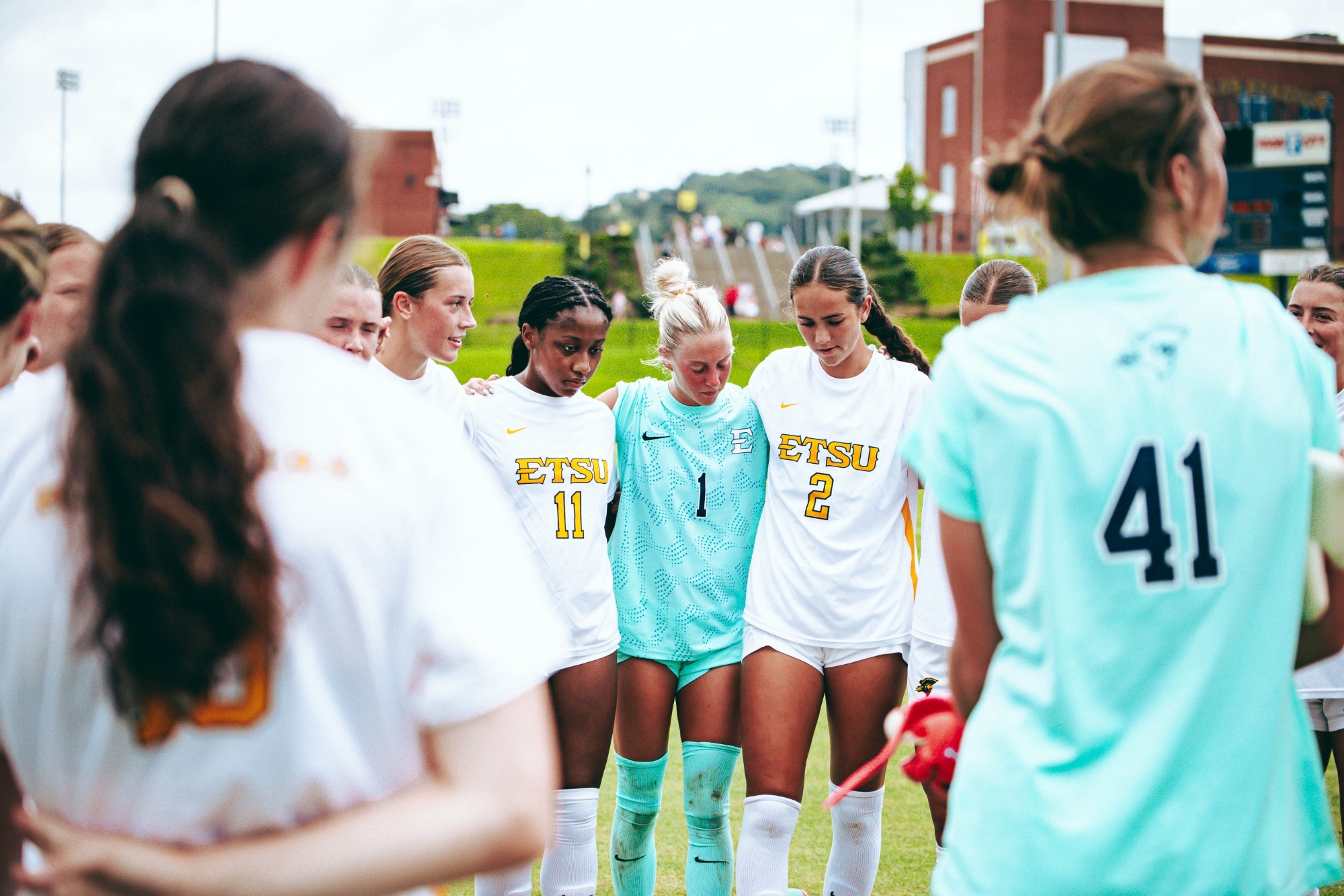 The ETSU women's soccer team huddles before a match with USC Upstate. The team is wearing their white uniforms except for the goalkeepers wearing teal. Jada Cooper, Finley Dybik, and Alex Beck are in focus within the huddle.