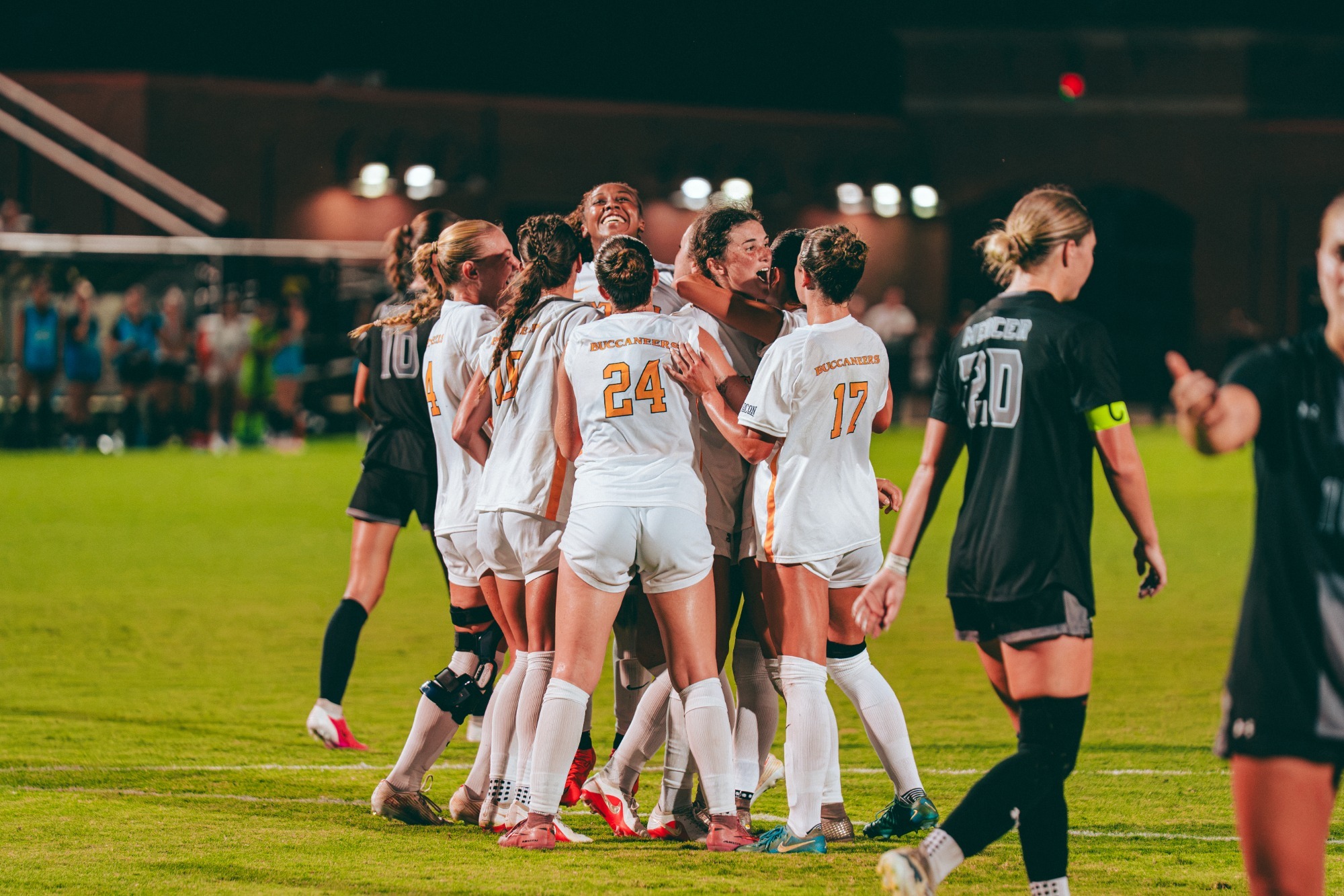 Soccer players in white jerseys with gold numbers surround Reagan Fitzgerald as she celebrates a penalty kick goal in a match against Mercer. A dejected Mercer player walks through the foreground.