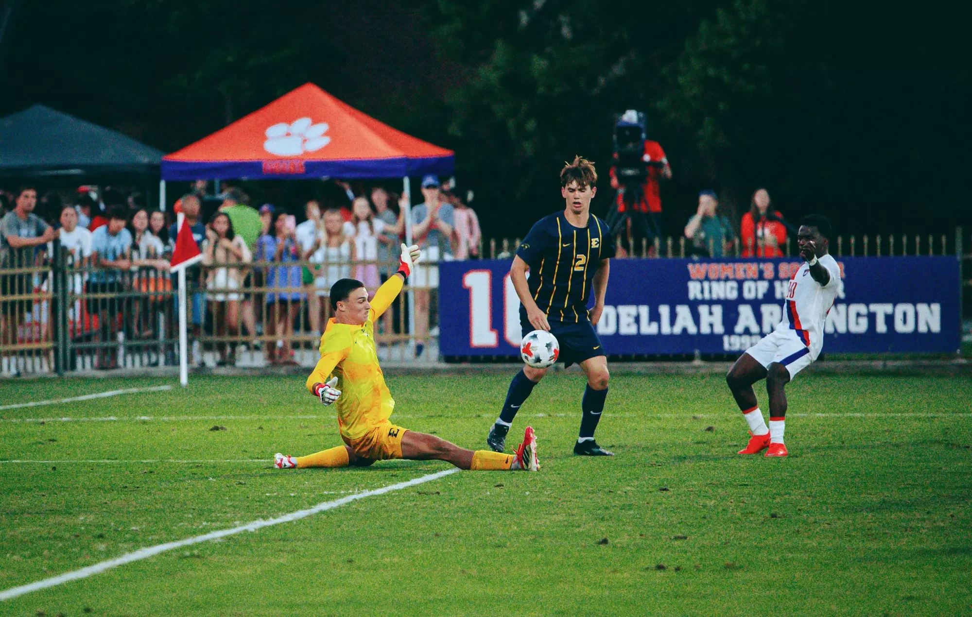 Noé Delmotte makes a sprawling save with his right hand while doing the splits during a soccer match against Clemson. Frank Manrod and a Clemson forward watch the ball as it approaches Delmotte.