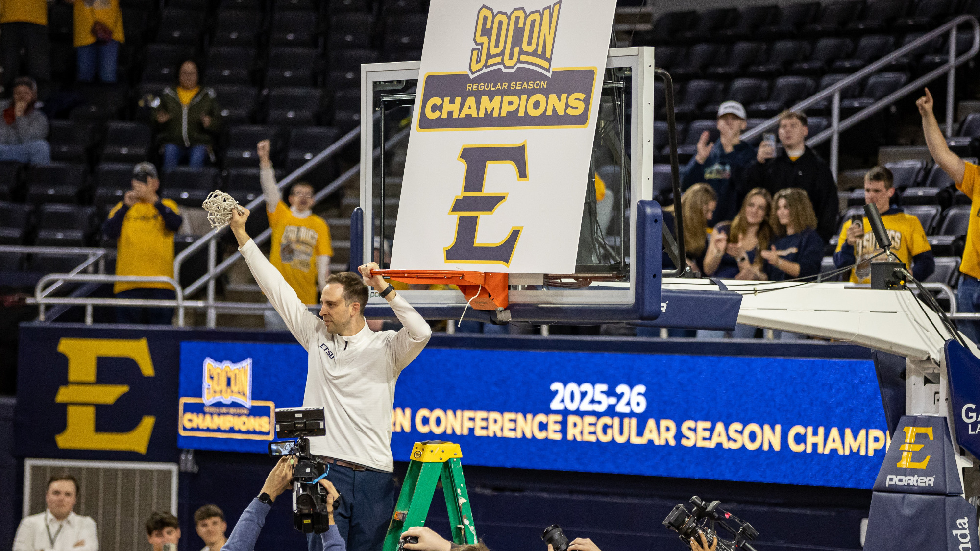 Brooks Savage holds up the remains of a basketball net while stadning atop a ladder. Behind him, a basketball goal supports a sign that reads 