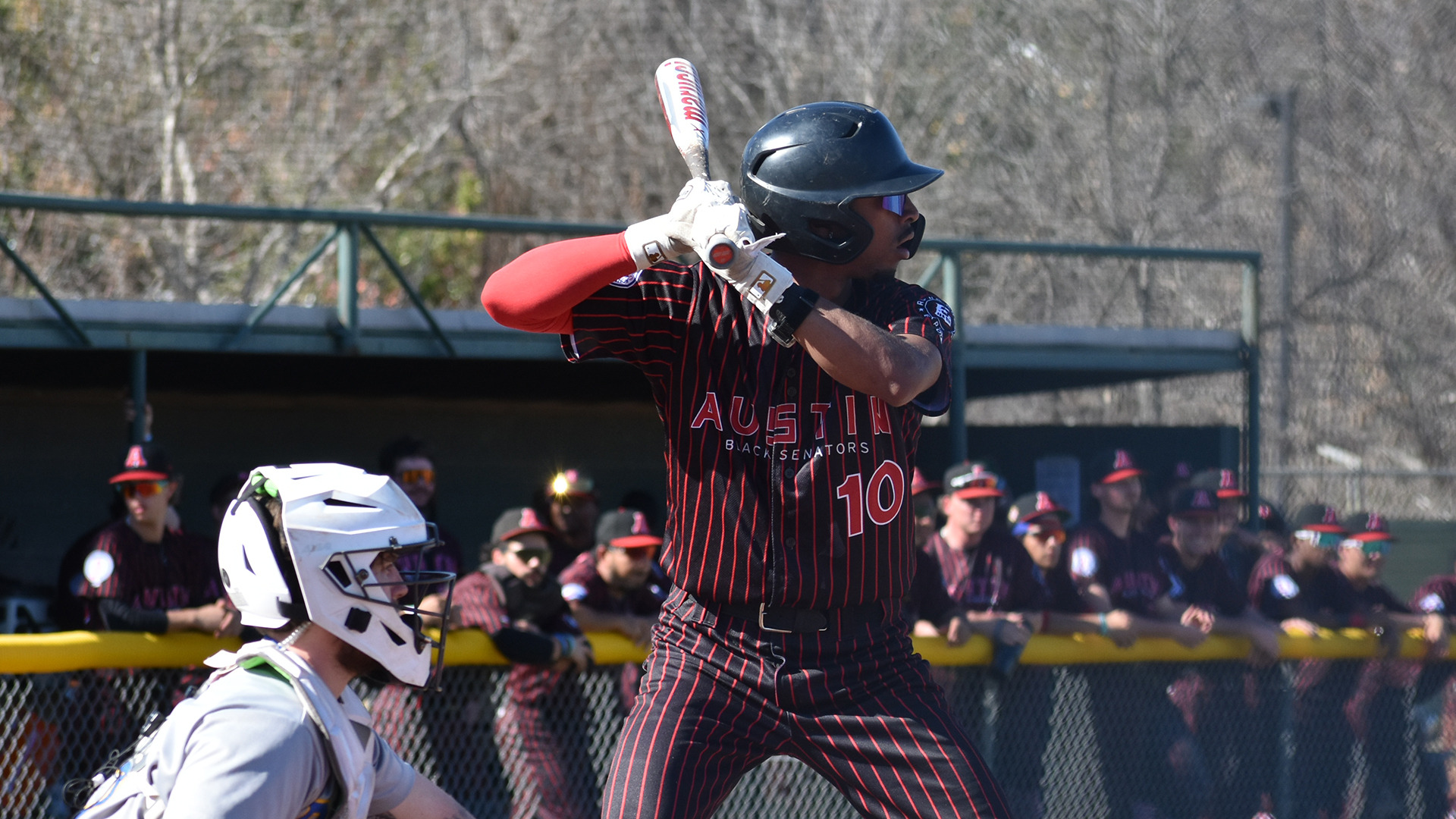 HT baseball player Jaden Corzine waits for a pitch at bat against Central Christian College. HT's dugout and players are visible in the background.