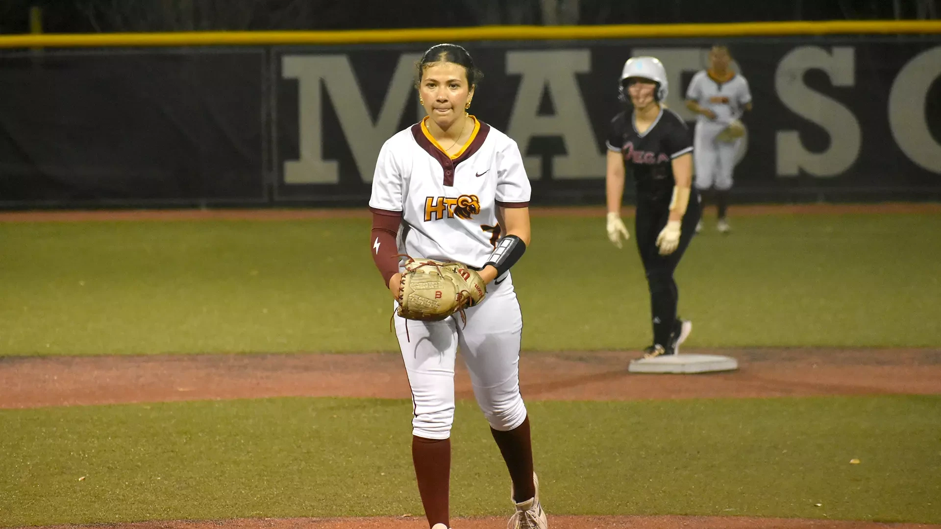 HT softball pitcher Alexis Tamez in the pitching circle about to wind up and throw a pitch. A Talladega baserunner is visible in the background on second base.