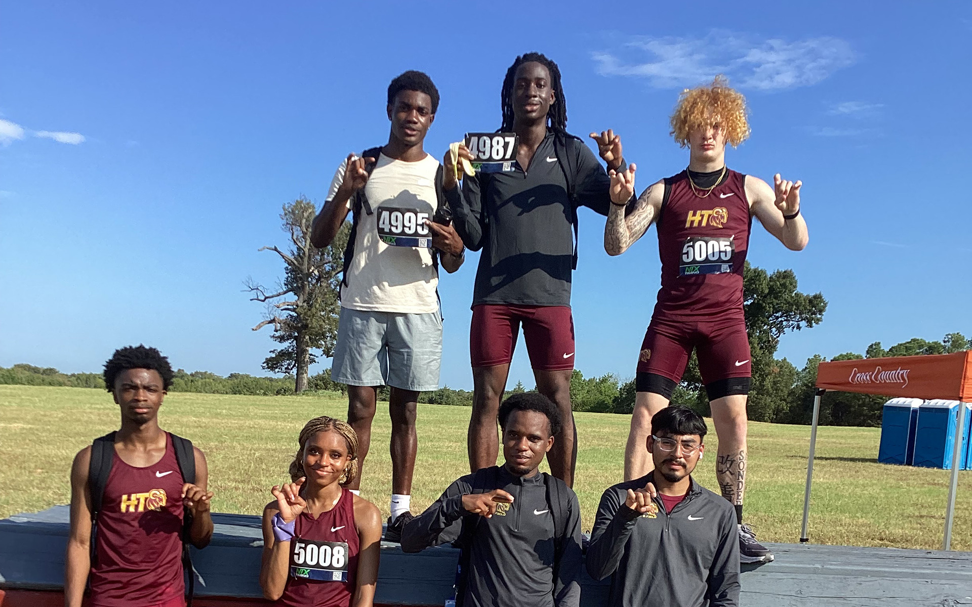 Six members of HT's men's cross country team and one member of HT's women's cross country team flash the Ram-horns hand signals after a meet in Tyler, Texas. Three men are on a platform at the top, and the remaining four competitors are on the bottom
