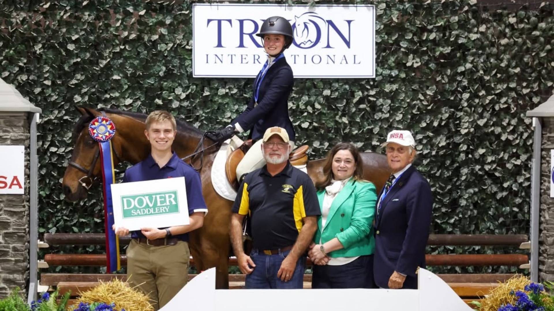 A group of five people and a horse pose at an equestrian event, holding awards in front of signs