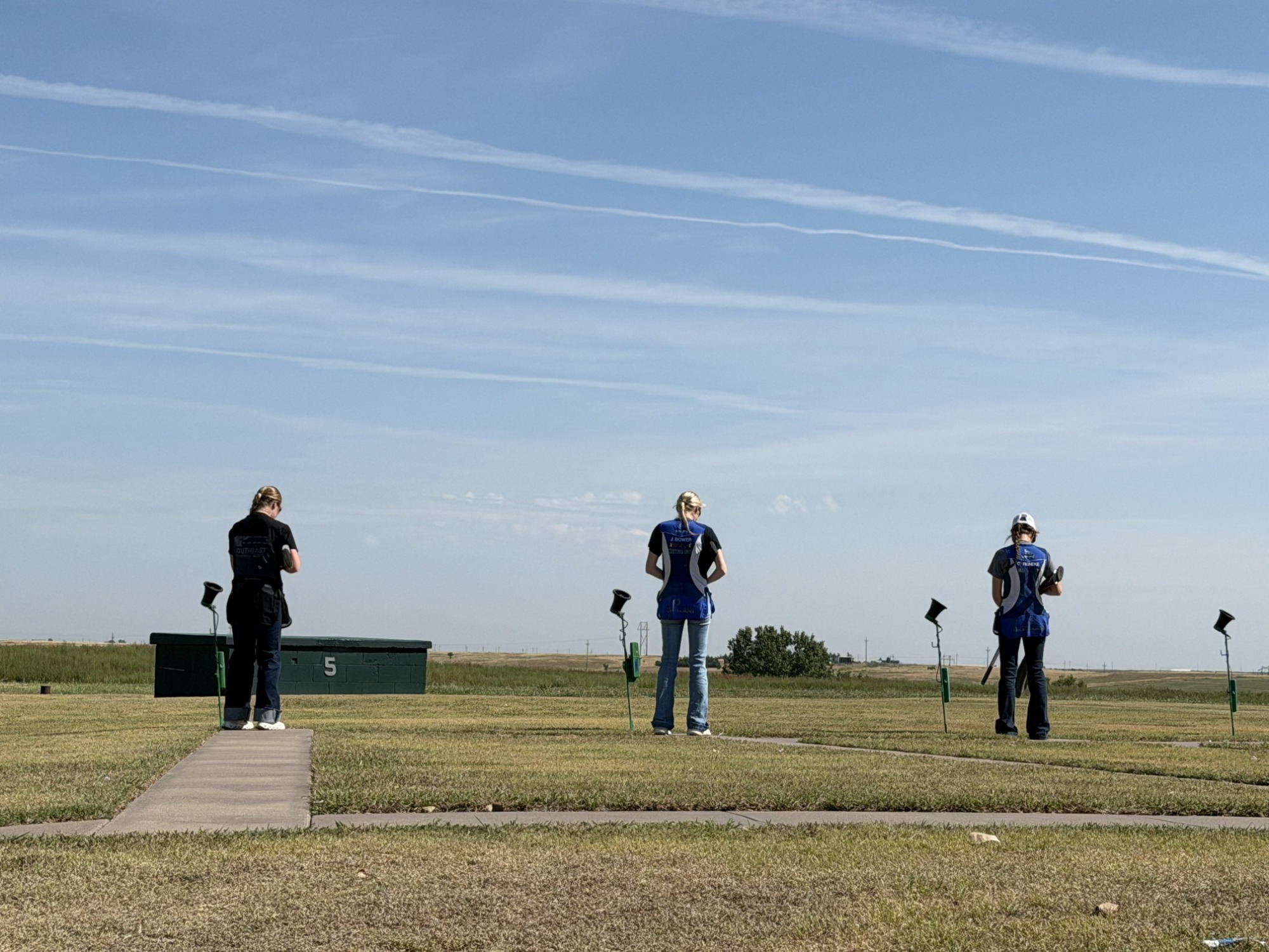 Three members of the SCC Shooting team stand lined up facing away from the camera and towards the throwers during an event, with the sky taking up most of the frame.